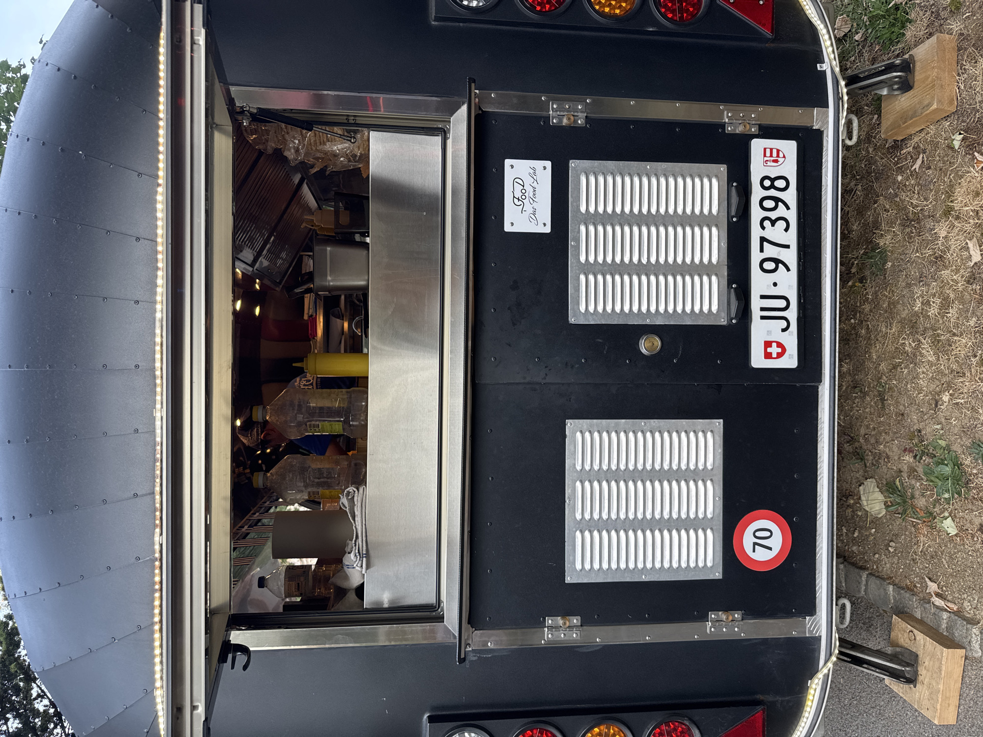 The image shows the rear of a black food truck parked outdoors on a slightly uneven, dirt and gravel surface. A portion of a tree is visible at the top left of the frame. The food truck has a service window that is open, revealing items inside, including bottles of liquid, paper towels, a metal container, and a yellow squeeze bottle. Below the service window are two black doors with silver ventilation grates on each. A circular red and white sign with the number "70" is affixed to the left door, indicating a speed limit. A rectangular Swiss license plate with the characters "JU - 97398" is mounted on the lower right side of the doors. A small white plaque with the text "FOOD Das Food Lab" is located on the right door above the ventilation grate. The food truck is supported by two metal legs with wooden blocks underneath. Several red and clear lights are visible on the exterior of the food truck on the far left and right edges of the image. The scene appears to be in a relatively quiet, possibly rural or suburban area.