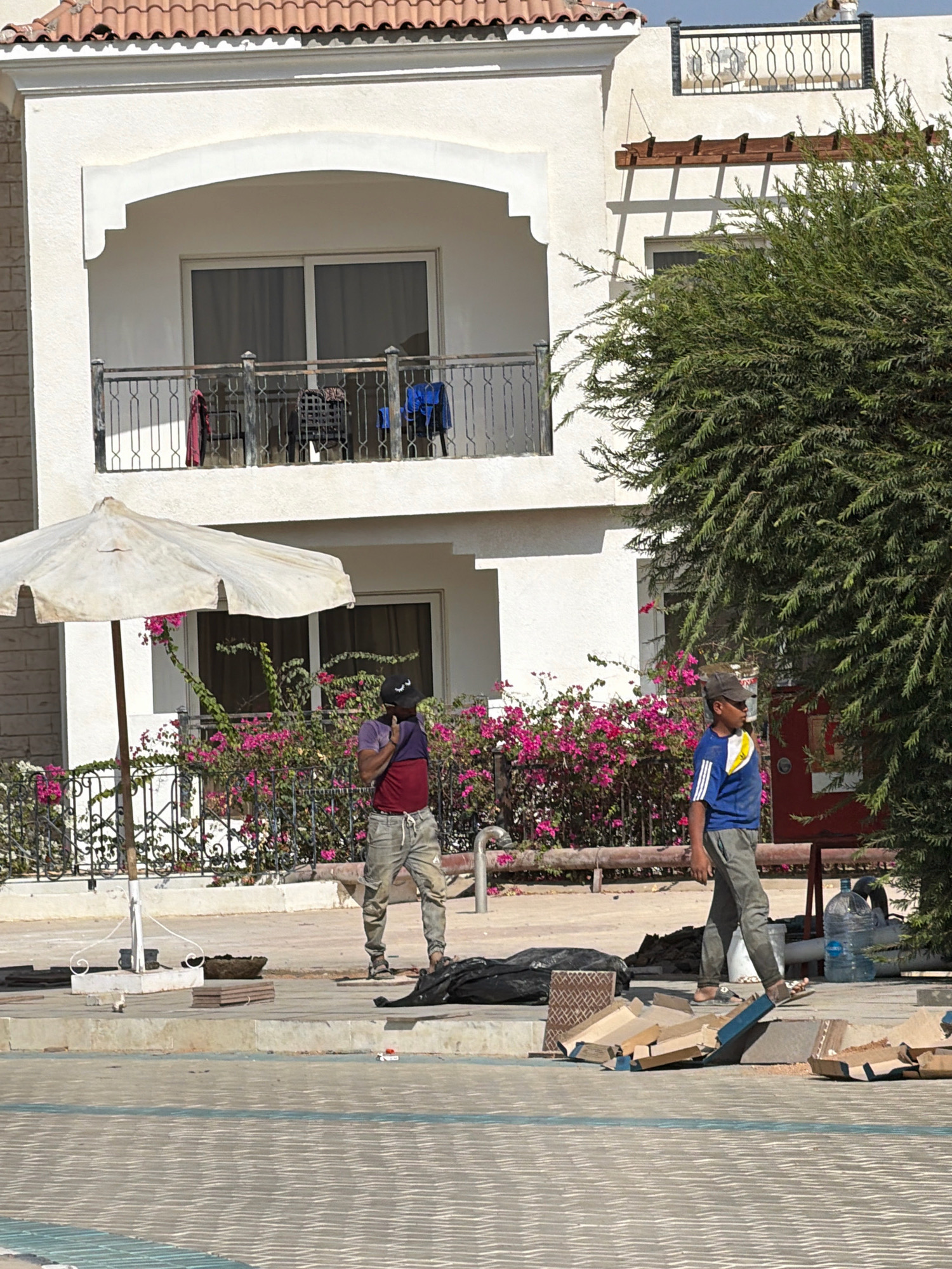 A scene unfolds on a sunny day in Sharm El Sheikh, Egypt, in front of a multi-story building with a tiled roof and white facade. The building features a balcony with a black metal railing, adorned with red and blue clothing and a black object. A large, white, weathered umbrella stands to the left, angled to provide shade. Behind it, a vibrant pink bougainvillea bush blooms.
Two individuals are present. The person on the left, wearing a purple and red t-shirt and light-colored pants, stands near the umbrella, holding their head with one hand and their neck with the other, as if shielding themselves from the sun or adjusting something on their head. They are also wearing a black baseball cap. To the right, a person in a blue t-shirt with yellow and white trim and grey pants walks away from the viewer. This individual is also wearing a dark baseball cap and appears to be wearing sandals.
The foreground of the scene is a patterned pavement, possibly a poolside area. Scattered on the ground near the individuals are construction-related materials: cardboard boxes, tiles, and a black trash bag. A large water bottle is visible behind the person in the blue shirt. The overall impression is one of an outdoor area, potentially a construction or renovation site, adjacent to a residential building. The bright sunlight suggests a warm climate.