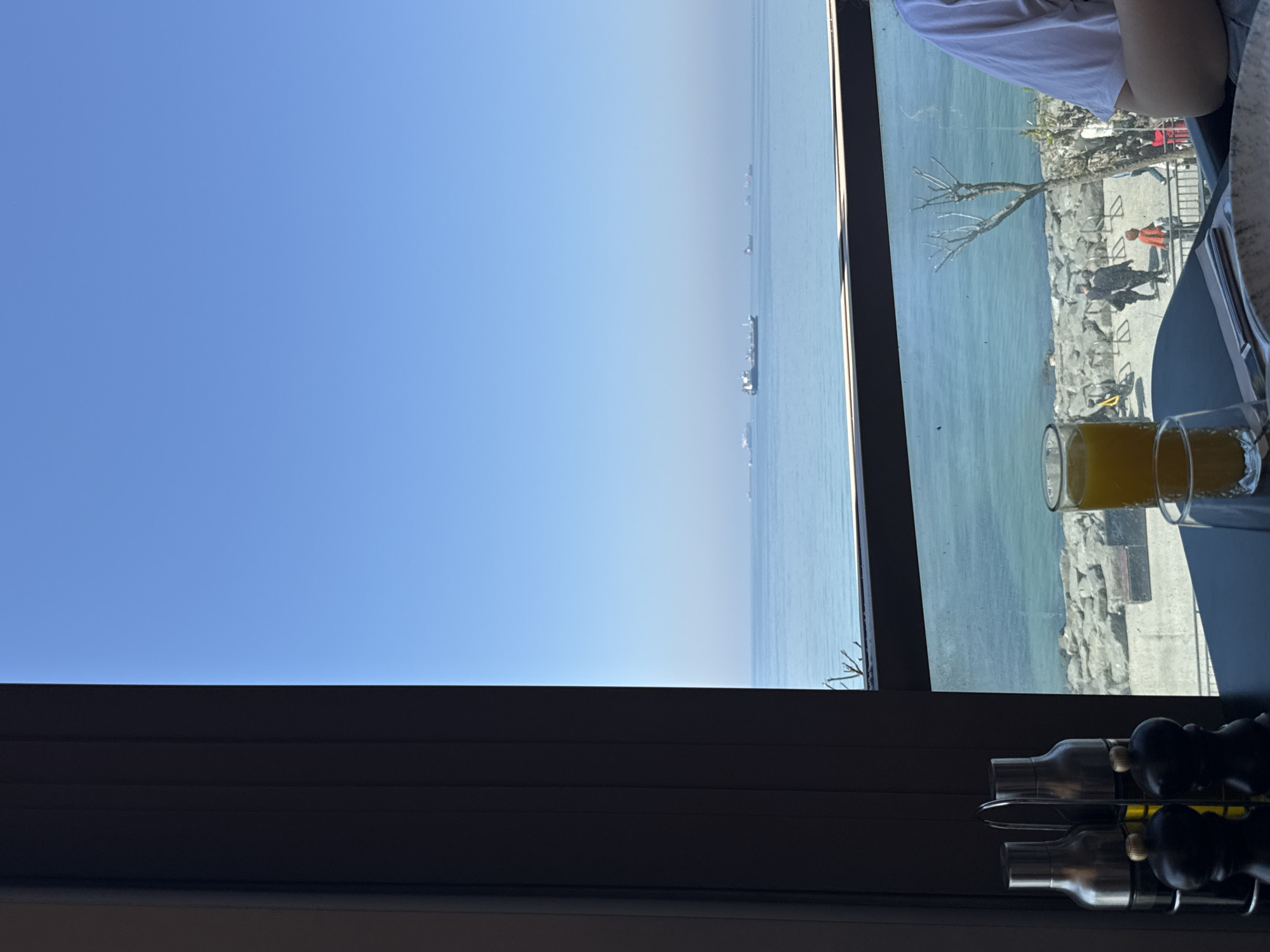 The image shows a view from a window. On the left, there is a clear blue sky with a thin horizon line. To the right, the scene transitions to a body of water. The window reflects a view of people walking along a rocky waterfront. In the lower right corner, there are glasses of orange liquid. Below the window, a metal rack holds seasoning containers. The location appears to be in a building in Bakırköy, Turkey, based on the visible landscape and characteristics of the water and sky.