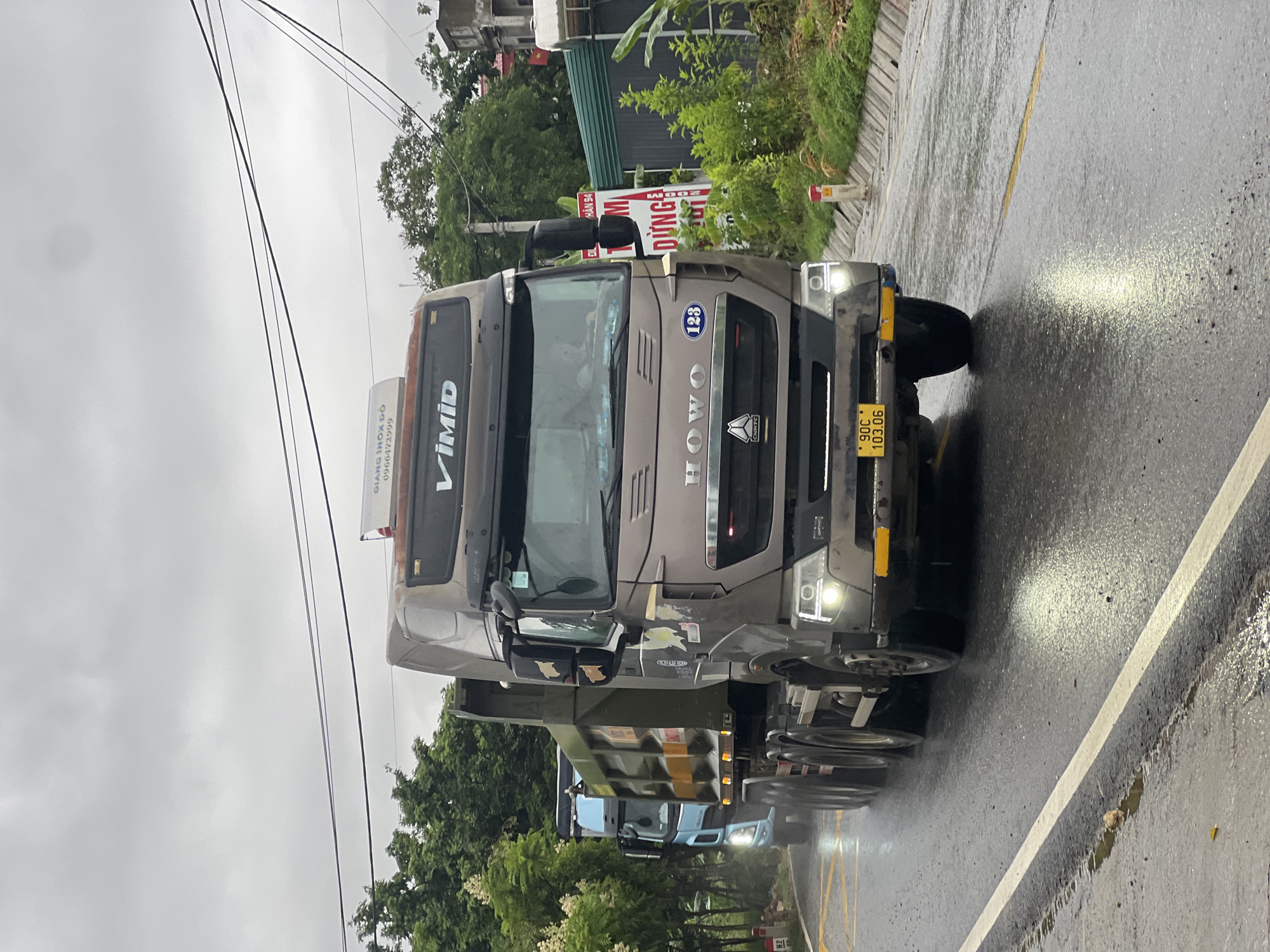 The image captures a rainy day scene on a paved road in Sơn Dương Commune, Vietnam. A large, brown dump truck is prominently featured in the foreground, driving on the right side of the road. The truck has the "HOWO" logo visible on its grille, and a license plate displaying "90C 103.06". Another blue truck is visible behind the brown one, also driving on the right. The road is wet, with reflections of headlights and the sky on its surface. Trees line the sides of the road, and buildings are visible in the background. The sky is overcast and grey, indicating a cloudy or stormy day. Electrical wires crisscross above the road. A red and white marker post is visible on the right side of the road. There are no visible people in the image.