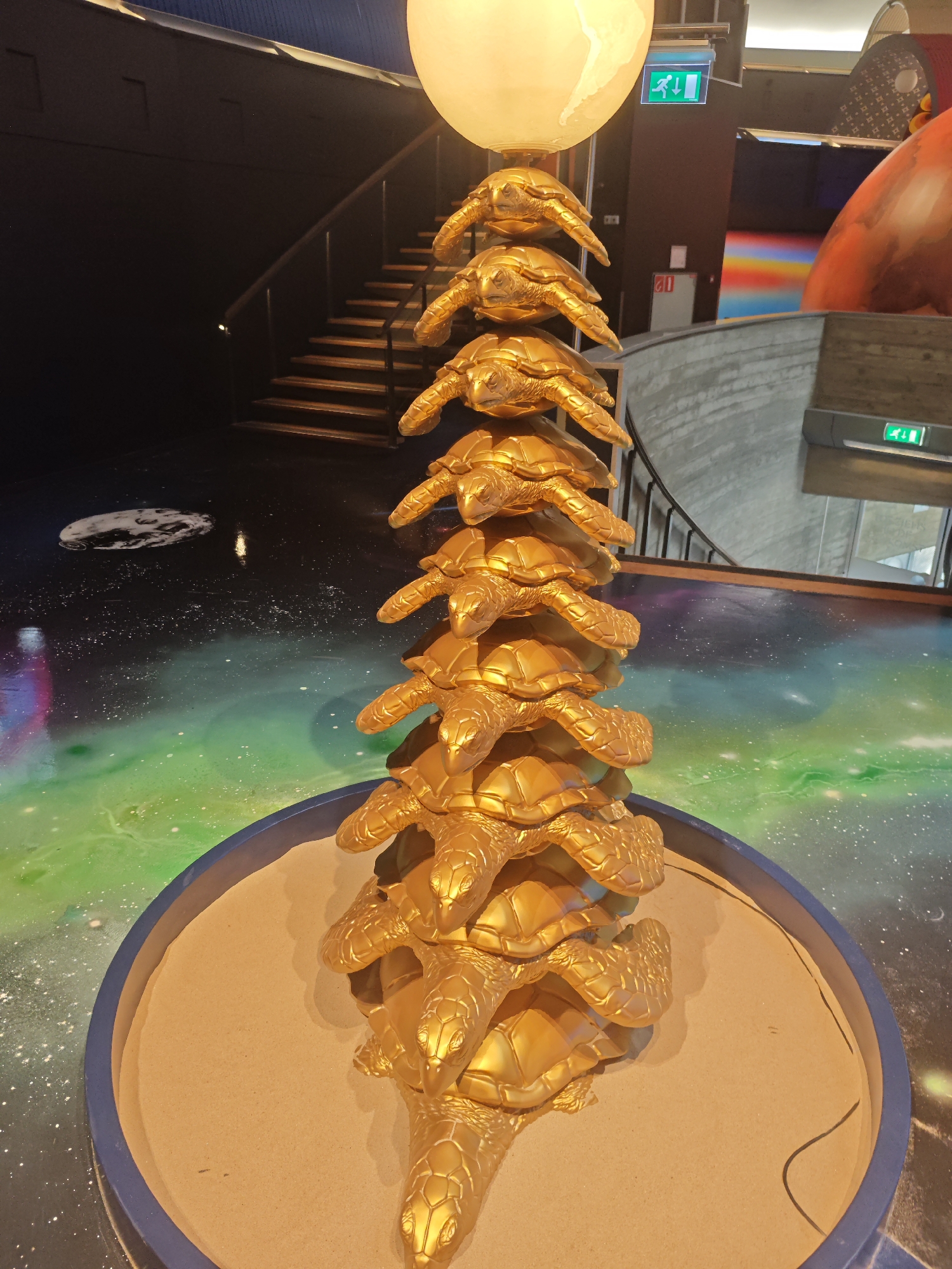 The image shows a golden sculpture of a tower of turtles, topped with a large, pale sphere, likely representing the Earth or the Moon. The sculpture is positioned in the center of a circular display filled with sand. The surrounding environment features a floor with a cosmic-themed mural depicting stars, nebulae in green and purple hues, and a circular representation of the Earth. In the background, a staircase with wooden steps and a dark railing is visible, leading upwards. An exit sign with a green running figure is mounted on a brown wall. A portion of a large, red, curved structure is also visible on the right side of the image. This scene is located inside the Evoluon in Eindhoven, Netherlands, a former science museum and conference center known for its distinctive UFO-shaped building and its current exhibition spaces. The lighting is dim, with the golden sculpture and the upper sphere being the brightest elements. There are no people visible in the image.