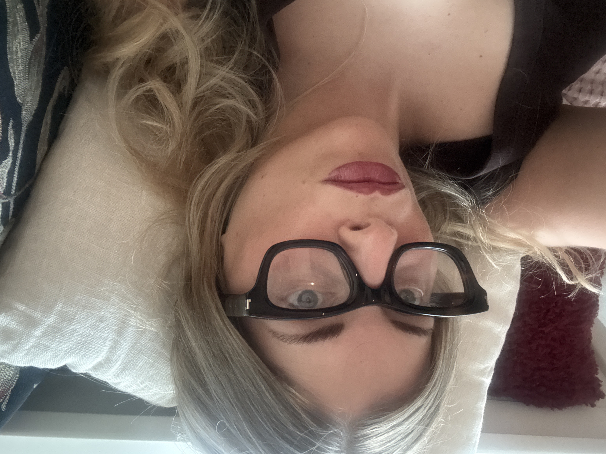 A woman with long, blonde hair and glasses is lying on a pillow. She has fair skin and is wearing a dark-colored top. Her lips are painted red. She appears to be indoors. There is no indication of the city of Paris, France.