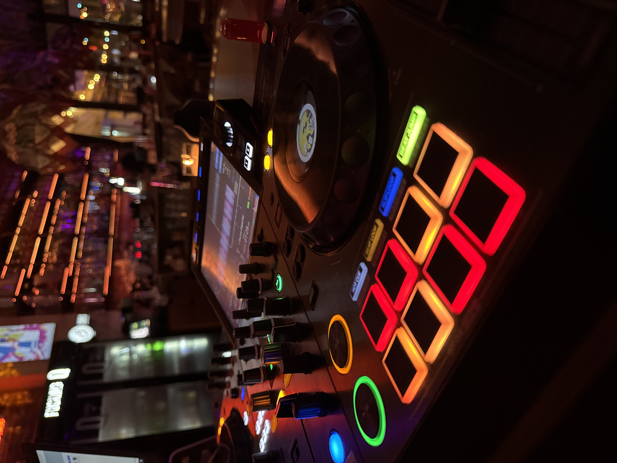 The image shows a DJ controller, likely a Pioneer DJ unit, with illuminated buttons and a screen displaying waveforms. The controller is surrounded by bar-like lighting. In the blurred background, there are reflections and lights suggestive of a bar or restaurant interior. Based on information embedded in the image metadata, the location is Plovdiv, Bulgaria.
