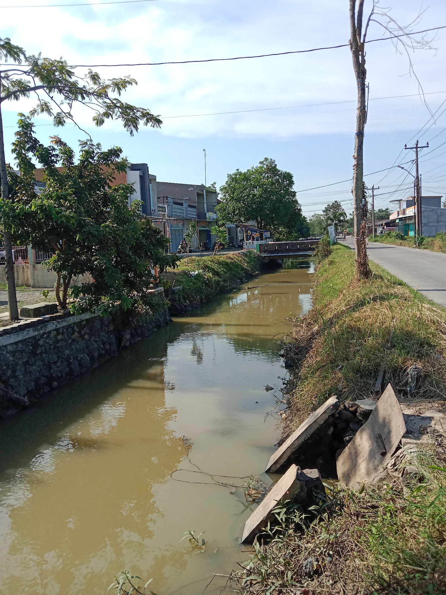 The image depicts a drainage canal in Cangkring, Indonesia. The canal is filled with muddy water. The banks of the canal are lined with vegetation and concrete structures. A small bridge crosses the canal in the distance. A road runs alongside the canal. Buildings and trees are visible in the background. Overhead power lines are present. Concrete debris is visible at the edge of the canal on the right side.