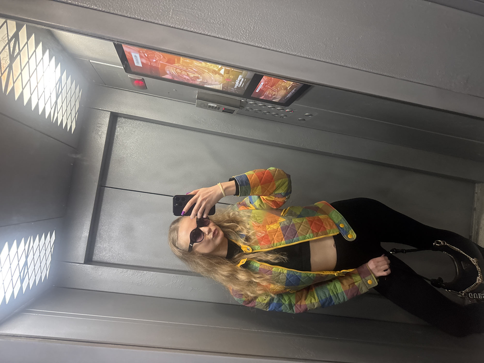 A woman is seen in an elevator taking a mirror selfie with her black smartphone. She is wearing a multi-colored quilted jacket with yellow trim over a black crop top and black leggings. She also has black sunglasses on. The elevator has gray metal walls and a ceiling with an illuminated grid pattern on the left and a panel with buttons and a display screen at the top. A Guess handbag can be seen in the lower right. The location is Unknown, Unknown.