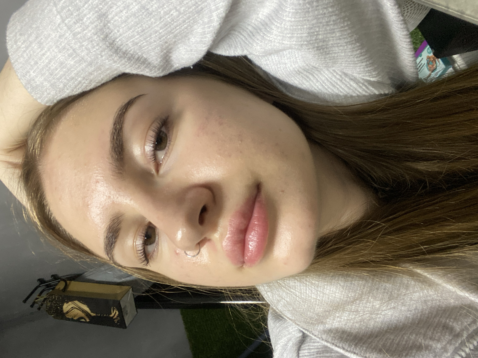 A medium close-up photograph depicts a young woman in a reclining posture, facing the camera directly. Her head rests on her right arm, which is covered by a light grey, textured, long-sleeved garment. She has light brown hair, visible flowing across her shoulders and down the left side of the frame. Her brown eyes are open, with long, dark eyelashes and well-defined eyebrows. Her skin exhibits a natural texture with some visible pores, minor blemishes, and a sheen on the forehead, nose, and chin. Her lips are full, pink, and appear glossy. A silver-colored hoop piercing is present in her left nostril. In the upper left background, a dark wall is visible, against which hangs a dark decorative item resembling a gift bag with a gold abstract design of a cocktail glass. Several thin, dark cylindrical objects are suspended from hooks near it. A small patch of green artificial turf is visible in the lower right background, behind her shoulder. Below the turf, blurred elements suggest product packaging, including a teal-colored box with orange details.