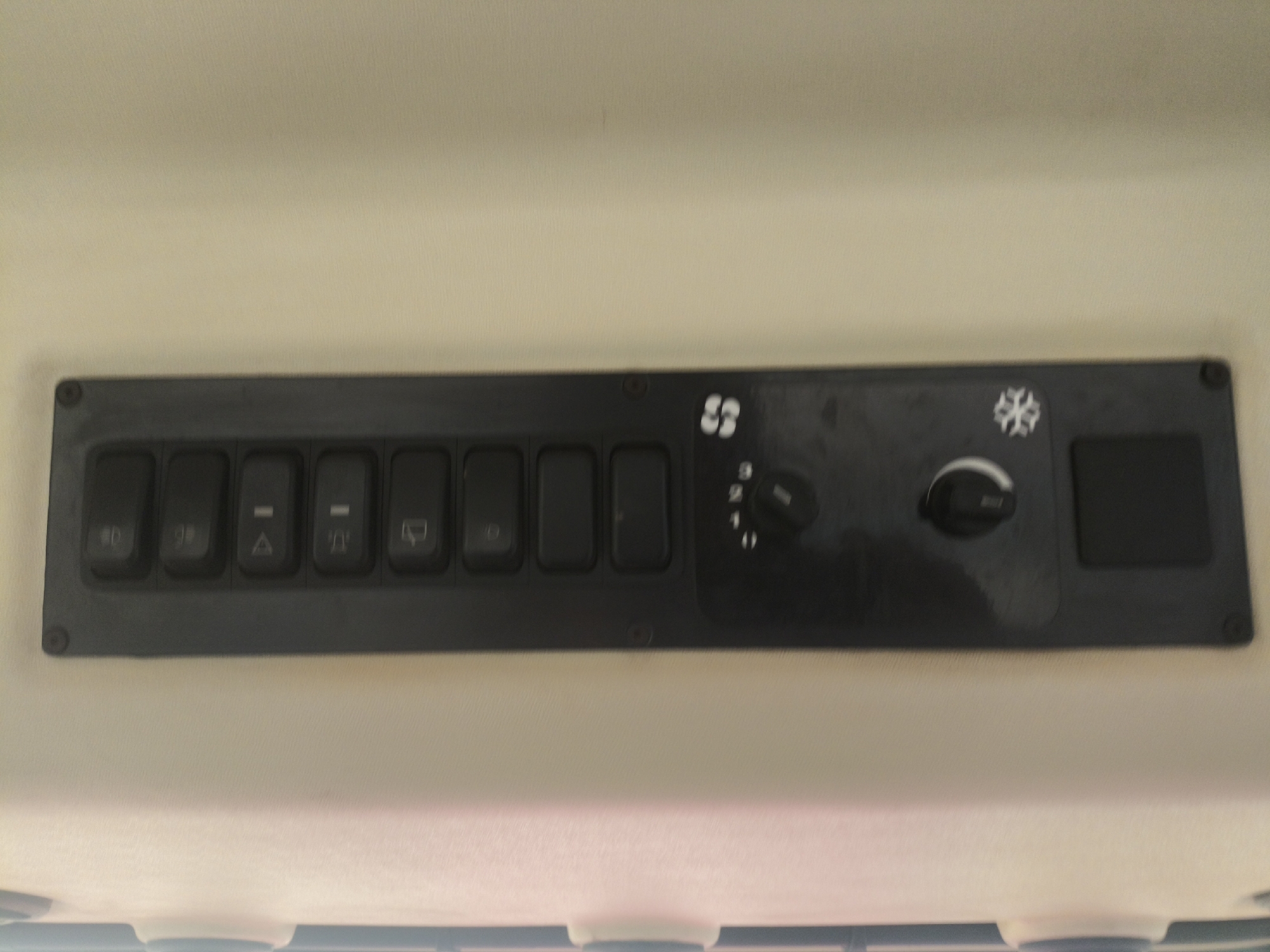 The image shows a close-up, eye-level shot of a control panel mounted on the ceiling of a vehicle, likely a tractor or construction equipment. The panel is black and rectangular, with several buttons and two rotary knobs. From left to right, there are six rectangular buttons. The first button has a symbol resembling a light bulb. The second button has a symbol that appears to be "90". The third button has a triangle with a line through it. The fourth button has a similar symbol to the third. The fifth button has a windshield wiper symbol. The sixth button is blank. To the right of the buttons, there is a fan symbol with an arrow, and a rotary knob with settings numbered 1, 2, and 3. Next to that is a snowflake symbol, followed by another rotary knob. Finally, on the far right, there is a square blank button. The control panel is attached to a light-colored, textured surface, possibly the interior headliner of the vehicle. The image does not depict any specific location in the city of Krylov, Russia, as it is a close-up of an indoor control panel.
