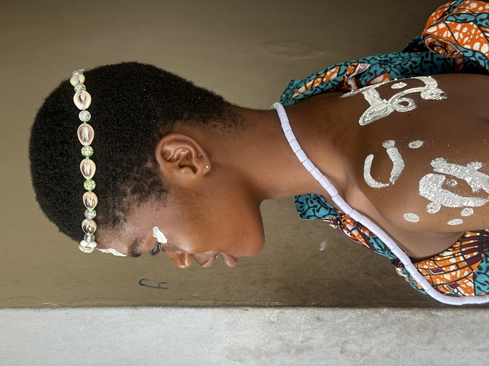 In Badagry, Nigeria, a young Black woman is shown in a profile view, looking to her left. She has short, dark, curly hair and a delicate golden stud in her left ear. A decorative headband adorns her forehead, featuring a row of small, light-colored shells interspersed with green beads. White markings are visible on her face, near her left eye, and on her shoulder and upper chest. She wears a white beaded necklace, and her shoulders are draped in a vibrantly patterned fabric with teal, orange, and white motifs. The background consists of a textured, light-colored wall on the left and a plain, muted brown wall on the right. A faint black mark, possibly graffiti, is visible on the light-colored wall.