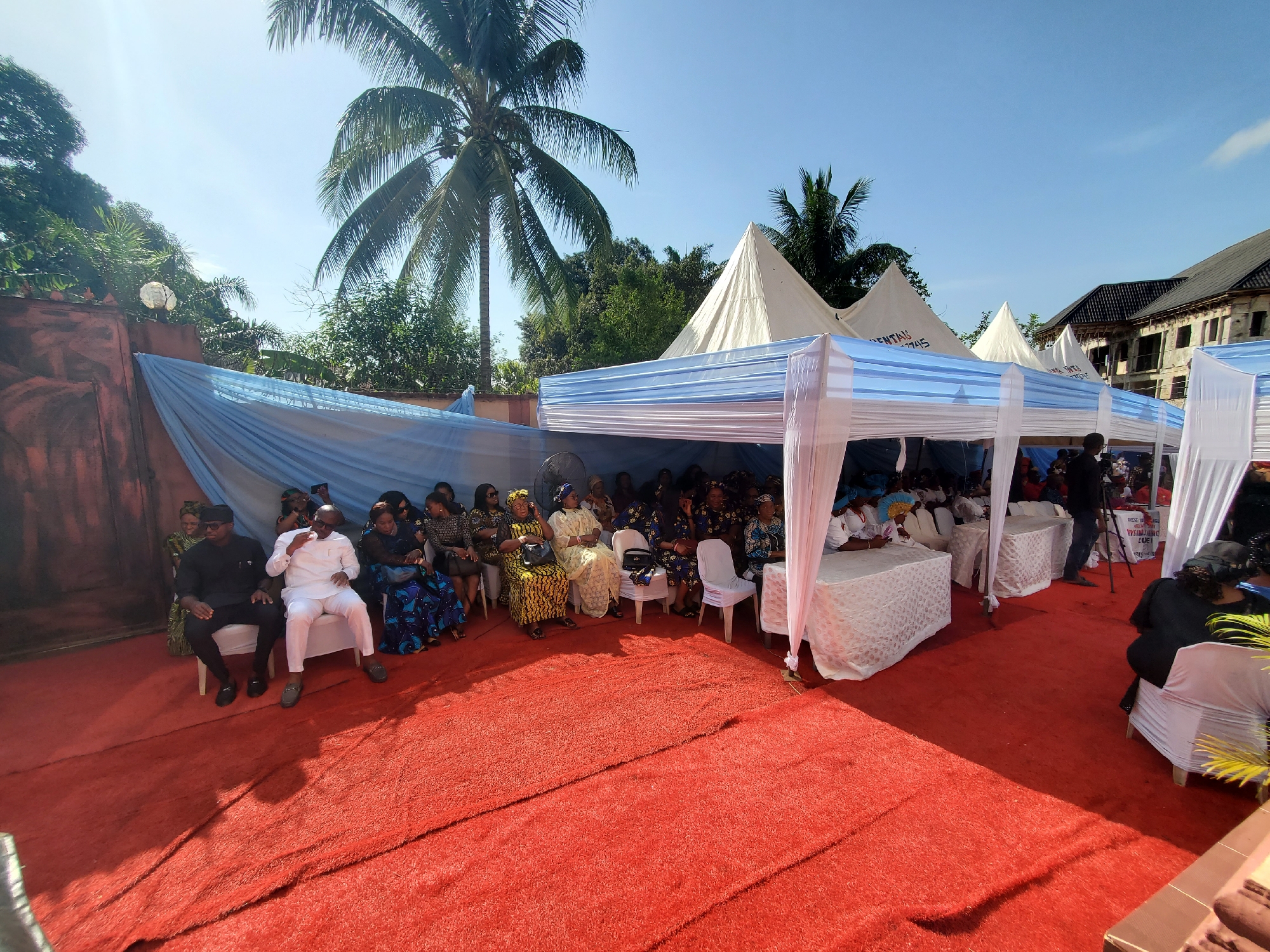 The image depicts an outdoor social gathering or event taking place in Obosi, Nigeria, under a bright, clear blue sky, suggesting mid-morning to early afternoon. The ground is covered with a vivid red artificial turf.
Numerous attendees, mostly adults, are seated on white chairs, some with white fabric covers, beneath a long canopy tent. This tent features white frames and a striped blue and white fabric roof, complemented by matching blue and white draped fabric along the sides. Many women are dressed in colorful traditional African attire, including patterned fabrics and headwraps, while some men wear formal black or white outfits.
In the foreground, individuals are seated, appearing attentive to the proceedings. Notable objects include small white tables with white tablecloths positioned under the tent. A standing fan is visible on the left side of the tent, providing comfort. In the background, beyond the main tent, several taller, white pyramidal tents are present. Partial text "RENTALS" and numbers like "7745" are faintly visible on one of these background tents. Another tent appears to show "NWAP TYRCE".
The setting is characterized by lush green tropical vegetation, including several prominent palm trees, which frame the scene. A brown wall or fence is seen on the left, and a partially constructed or renovated building with a dark roof is visible in the far background on the right. A tripod, likely for a camera or video recording, is positioned near the right side of the main tent. The overall scene suggests a well-organized temporary event space, likely for a celebration or ceremony, with a calm and orderly atmosphere.