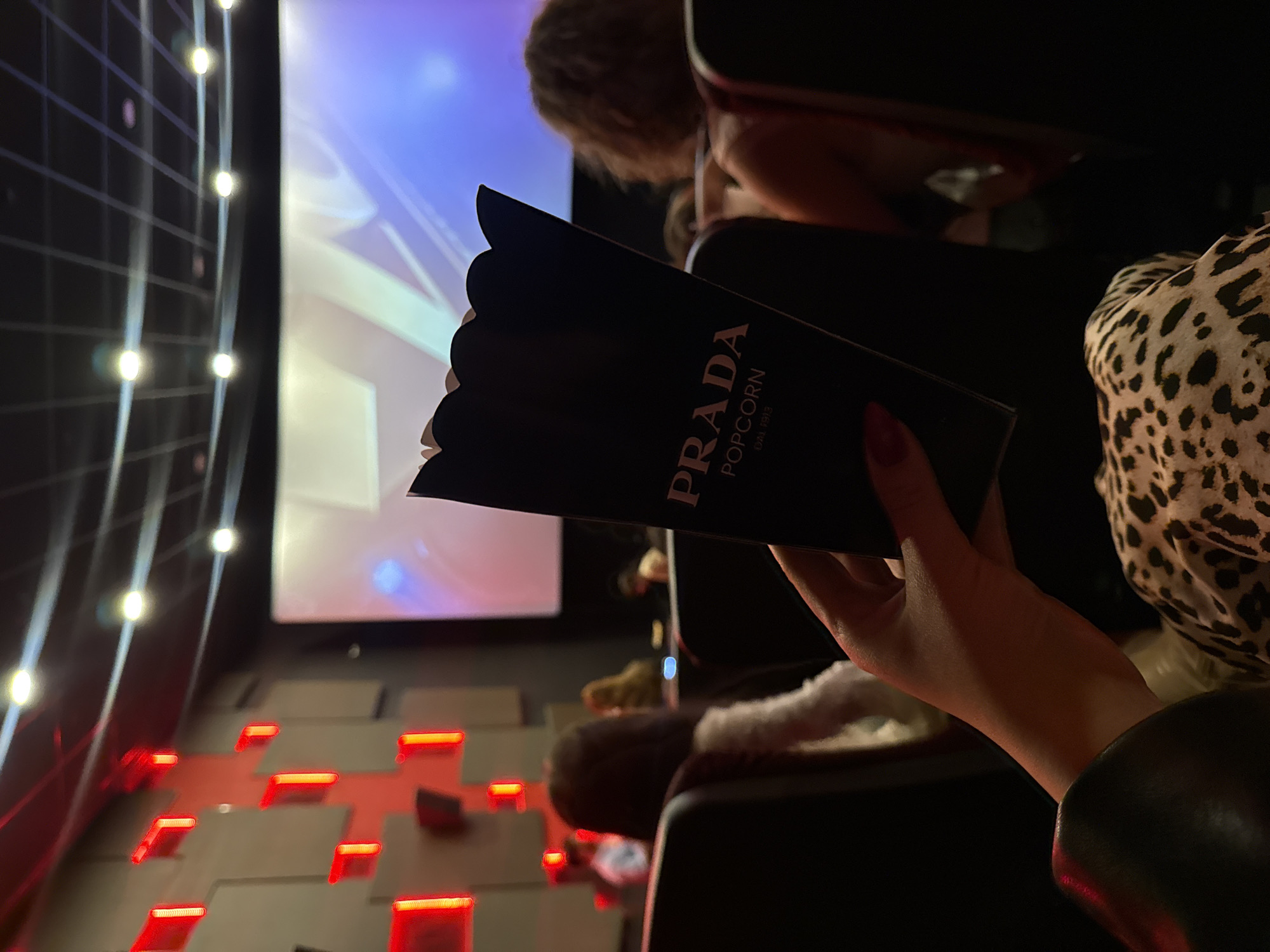 Here is a forensic description of the image:
The image shows the interior of a movie theater, potentially in Yerevan, Armenia, as indicated by the "Prada Popcorn Est. 1913" box, a collaboration between the brand and the Cinema Star multiplex located in Yerevan. The focus is on a black popcorn box held by a fair-skinned hand with red nail polish. "PRADA POPCORN Est. 1913" is printed on the box. The hand and box are positioned between rows of seats. An individual with short brown hair can be seen in the upper center of the frame. A person in an animal-print top sits to the right. A movie screen showing an indistinct image is visible in the background, along with decorative lighting on the walls. The theater's lighting is dim, creating a dark environment. The seats appear to be upholstered in black leather.