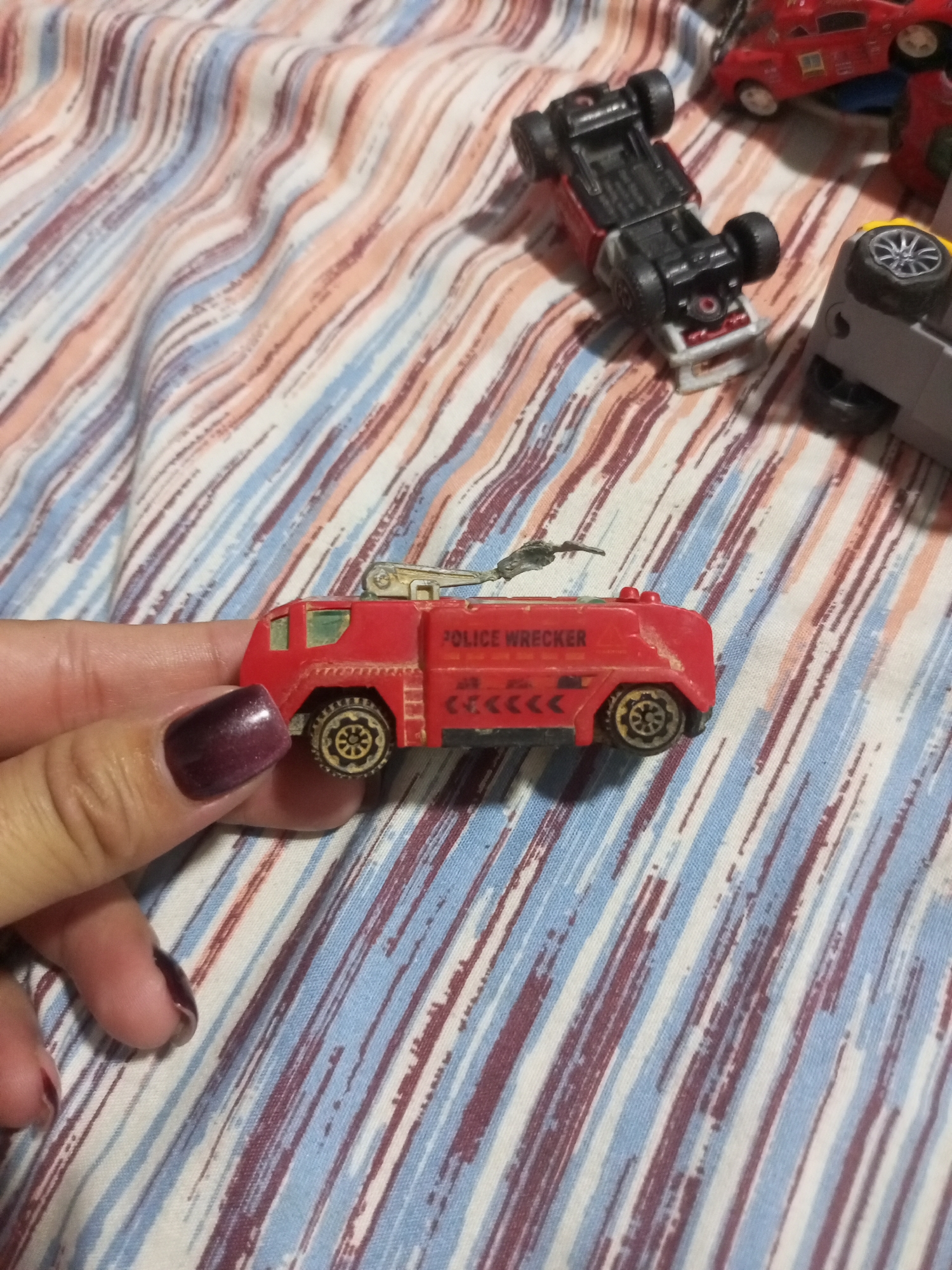 A close-up shot shows a toy red police wrecker truck being held by a hand with dark purple nail polish. The truck is positioned on a striped fabric, likely a bedspread, with diagonal lines of white, pink, and blue. The truck has "POLICE WRECKER" written on its side in yellow letters, and a silver crane-like apparatus on top. Other toy vehicles, including a red car flipped upside down and a grey vehicle also flipped over, are visible in the background on the same fabric. The scene is indoors, with no visible environmental context to place it in La Tagua, Venezuela.