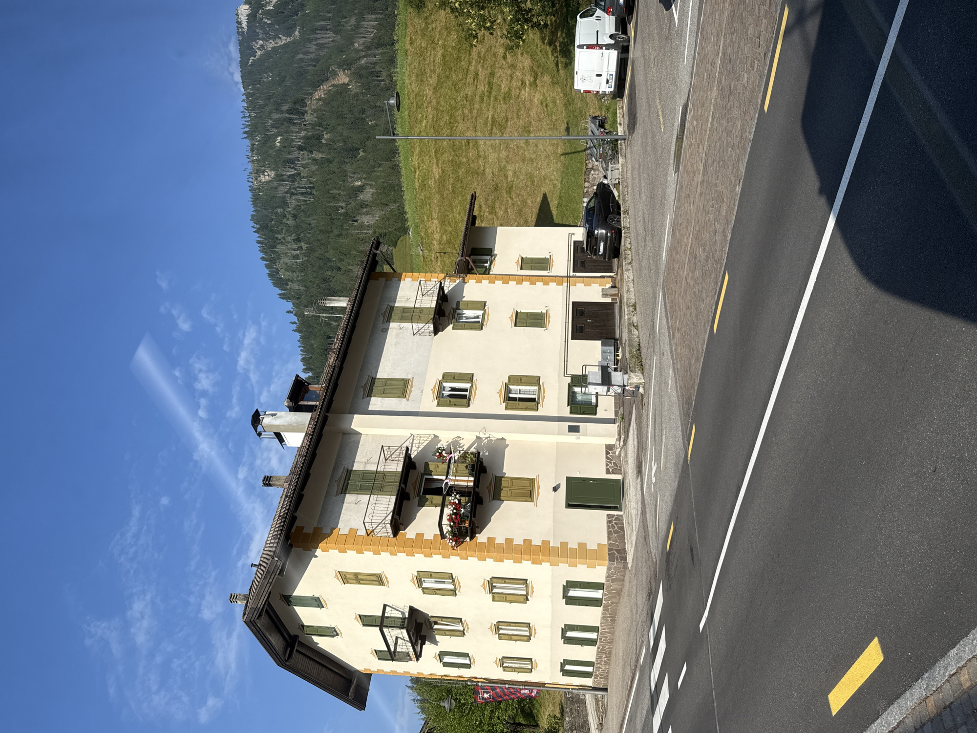 The image depicts a street scene in Moena, Italy, characterized by a building with cream-colored walls and yellow accents, featuring multiple windows with green shutters and balconies. A paved road with white and yellow markings runs in the foreground. Adjacent to the building is a parking area with several vehicles, including a dark SUV, a white van, and a black car. In the background, a steep, tree-covered mountainside rises under a clear blue sky with scattered clouds. The overall environment suggests a daytime setting in a town nestled within a mountainous region.