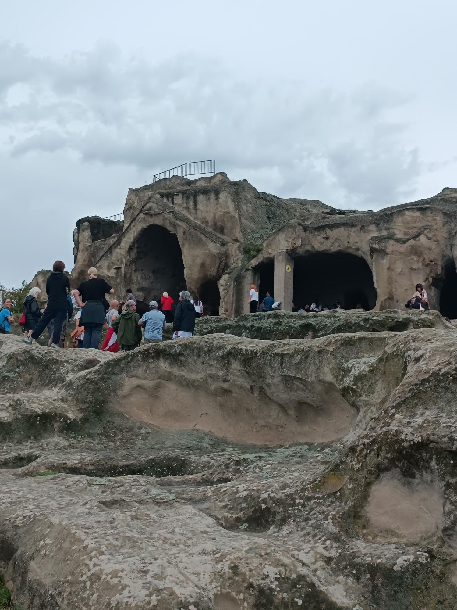 This is a wide, eye-level shot of the ancient cave city of Uplistsikhe, Georgia, on an overcast day. The foreground is dominated by the textured, light-brown rock formations that characterize the city. In the midground, a group of approximately 15 to 20 people are spread across the rock ledges and near the entrances of several large caves carved into the cliff face. The people are dressed in casual, everyday clothing and appear to be tourists observing the historical site. They are standing, sitting, and walking around, some pointing or looking into the caves. The cave entrances are dark, revealing only glimpses of the interiors. The background shows more of the cave city's structures, with a railing visible at the top of one of the formations, suggesting a viewing platform. The sky is a uniform, light gray, contributing to the somber mood of the scene.