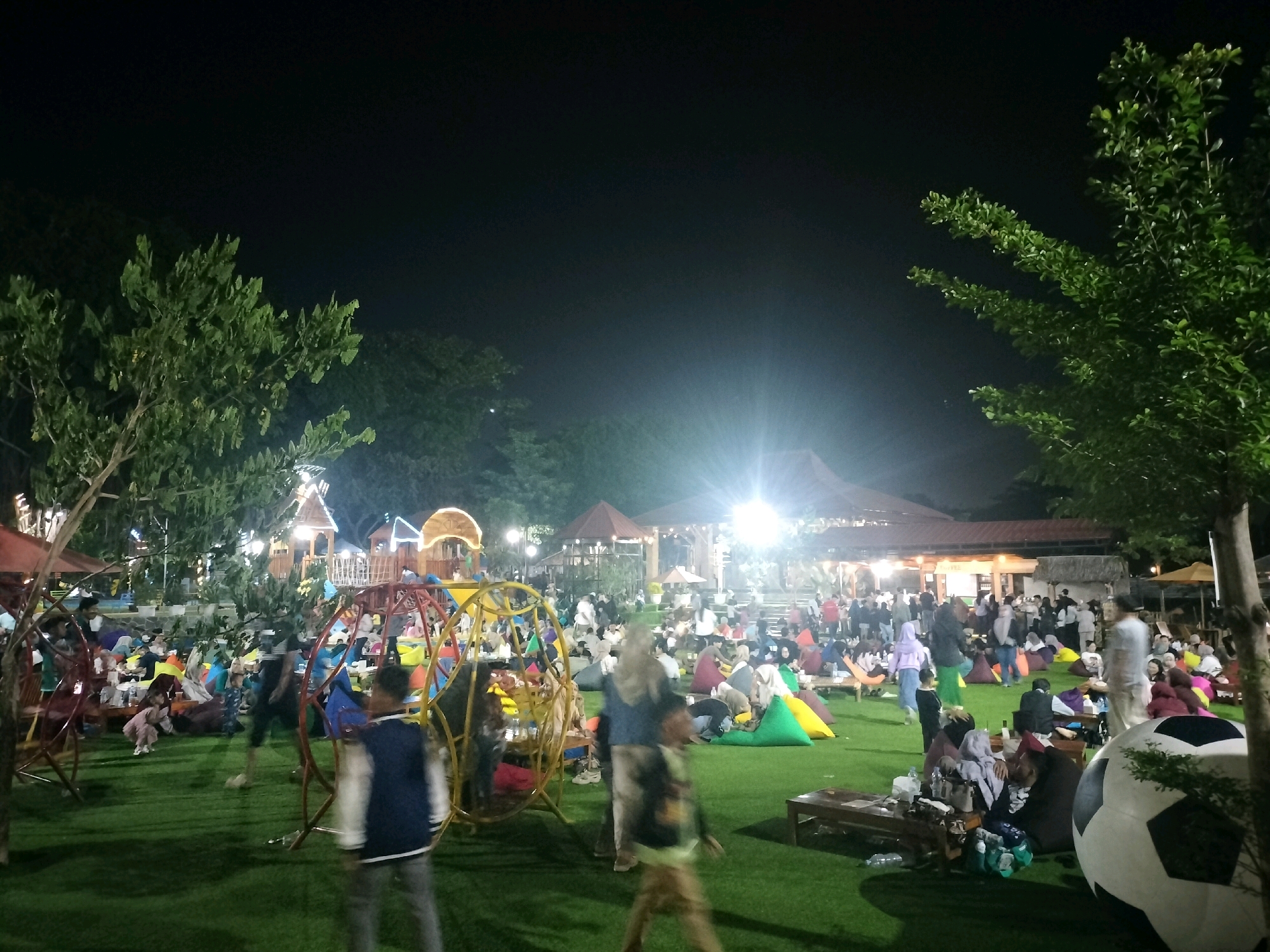 The image depicts a lively outdoor gathering at night in Depok, Indonesia, likely at a family-friendly entertainment venue or restaurant. The foreground features a bright green, artificial turf lawn scattered with large, colorful beanbag chairs. Several children are visible playing on a large, spherical, metal playground structure painted in vibrant colors of yellow, red, and blue. In the mid-ground, a crowd of people, including families and individuals, are seated on beanbags and at low tables, enjoying food and drinks. Numerous wooden structures, possibly stalls or seating areas, are visible in the background, illuminated by warm, bright lights, creating a festive atmosphere. A prominent, large, black and white soccer ball decoration is situated on the right side of the frame, adding to the recreational theme. The overall scene is one of a casual, enjoyable evening out, with families and friends relaxing and interacting in a well-lit, communal space. The presence of trees and foliage indicates an outdoor setting.