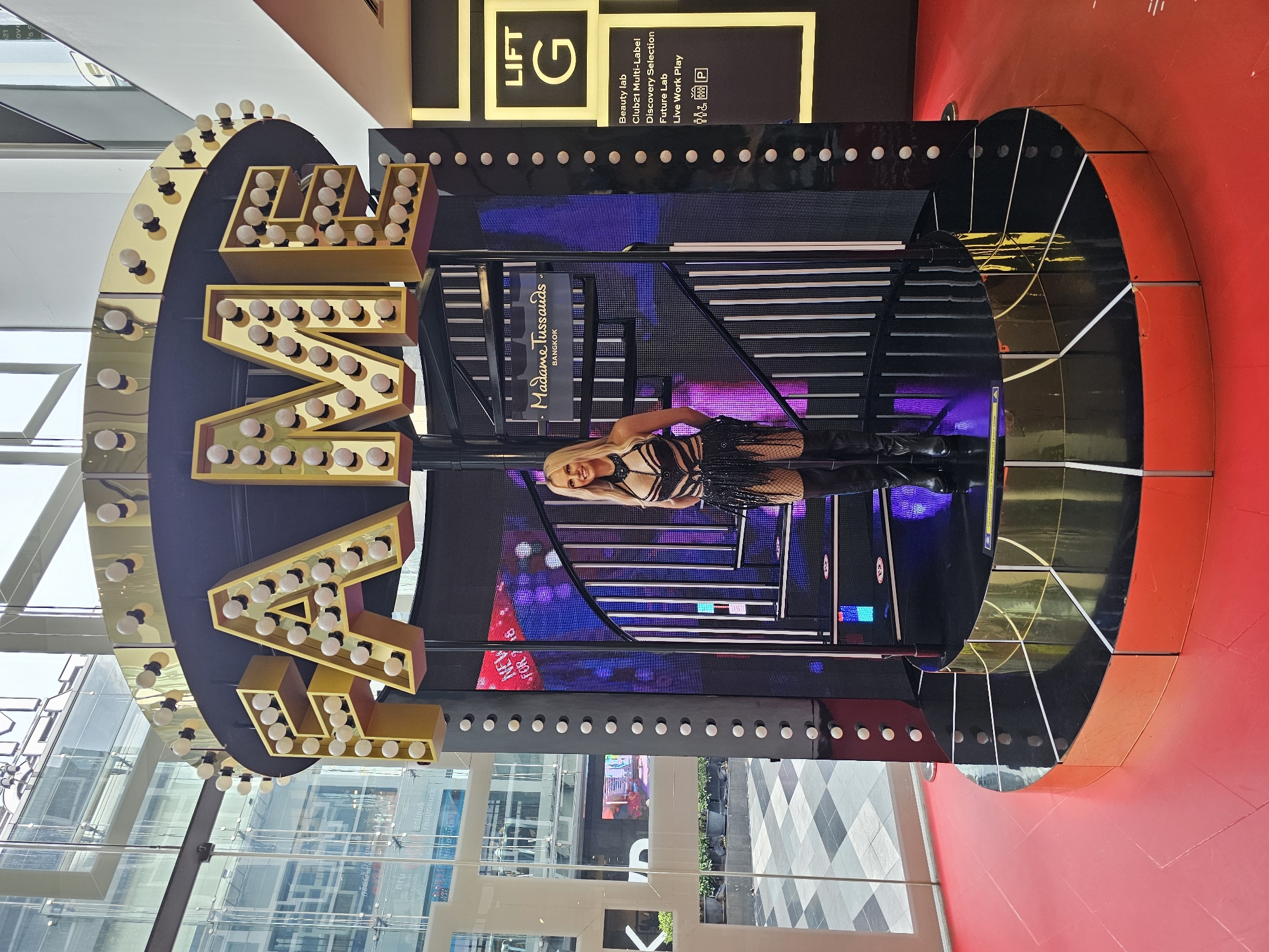 The image shows an outdoor display in Bangkok, Thailand, featuring a Madame Tussauds exhibit. The central focus is a wax figure of a woman dressed in a black, revealing outfit with fringe detailing and thigh-high black boots. She stands on a multi-tiered, circular stage with a black reflective surface and a gold trim base. Behind her is a spiral staircase that leads up to a large, illuminated sign spelling out "FAME" in golden, bulb-encrusted letters. The background includes a glass-fronted building, suggesting an indoor-outdoor shopping mall or public space. To the right of the exhibit, a black panel displays the text "LIFT G" above a list of labels including "Beauty lab," "Club21 Multi-Label," "Discovery Selection," "Future Lab," and "Live Work Play," along with icons representing men, women, a wheelchair, and a parking symbol. The floor around the exhibit is bright red. The overall scene is designed to evoke a sense of celebrity and glamour.