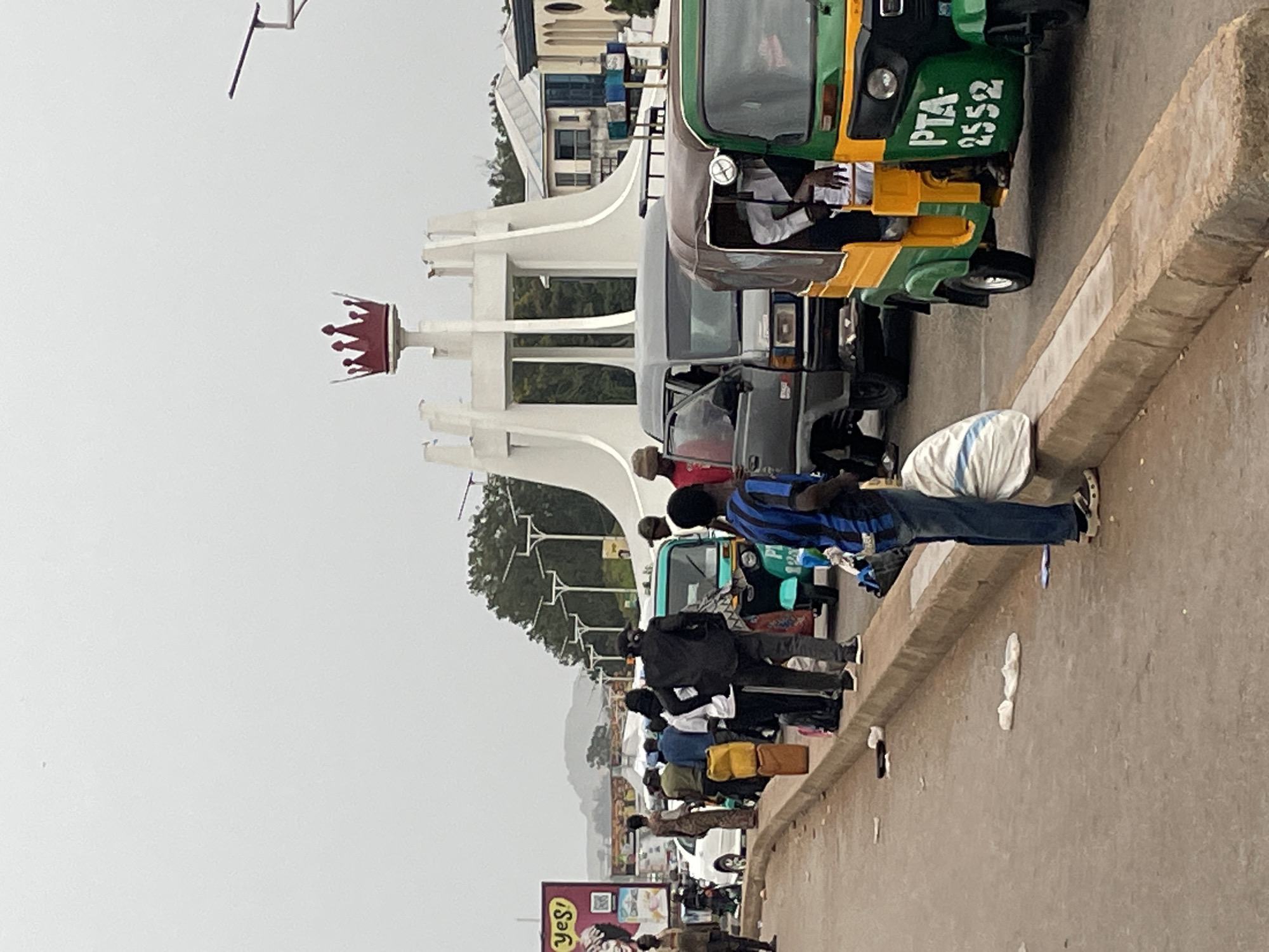 The image depicts a street scene in Jos, Nigeria, near a prominent monument featuring a large red crown. The monument is white and has a modern, abstract design with pillars. In the foreground, a line of people is gathered by the roadside, some carrying bags and luggage, suggesting they are waiting for transportation. Several vehicles are present, including a green and yellow tricycle (a common mode of transport in Nigeria), a grey van, and a white car further down the road. The road is paved, and a concrete curb separates it from a sidewalk area where some of the people are standing. There is visible litter on the ground. A large billboard with the word "yes!" and images of people is visible in the background on the left. The sky is overcast.