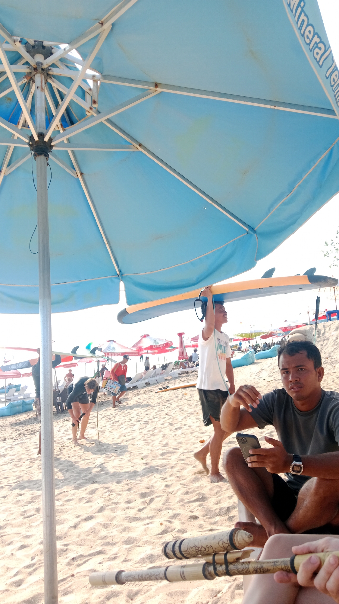 The image depicts a sunny beach scene under a large blue umbrella. In the foreground, a man is seated and looking at his phone, with two decorative bamboo sticks resting in his lap. To his left, another man is walking away, carrying a surfboard over his head. Further in the background, several people are visible on the sand, some near beach chairs and umbrellas, suggesting a relaxed atmosphere. The beach appears to be populated with a few surfboards and other beach equipment. The environment is sandy, and the sky is bright and clear, indicating a warm day.