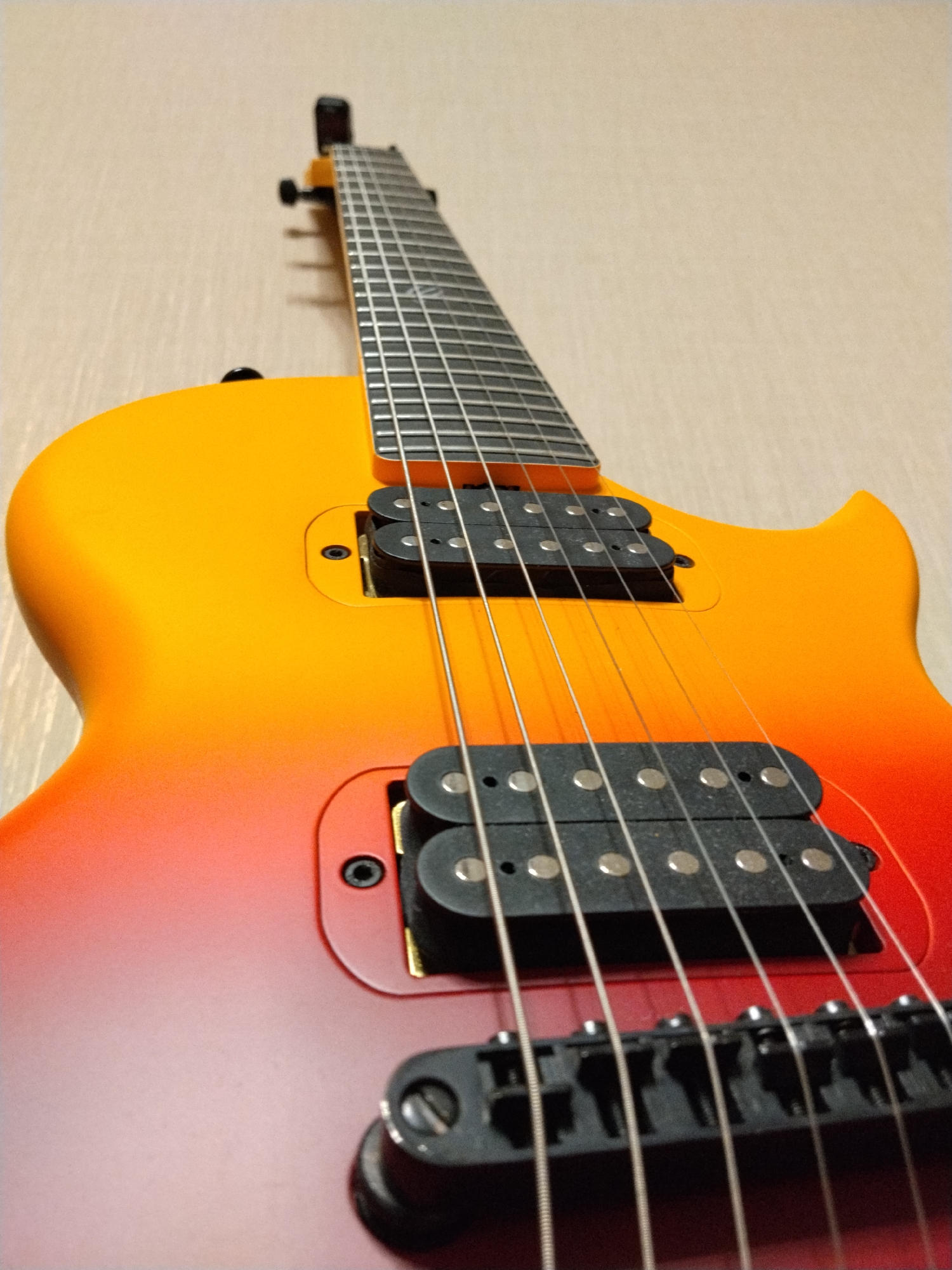 A close-up, low-angle shot shows the body and neck of an electric guitar. The guitar has a sunburst finish, transitioning from orange at the top to red at the bottom. Two black humbucker pickups are visible on the body, along with the strings and bridge. The guitar is leaning against a light-colored, textured wall, possibly wallpaper. The image is shot in what appears to be an indoor setting. No specific location in Krasnogorsk, Russia is evident.