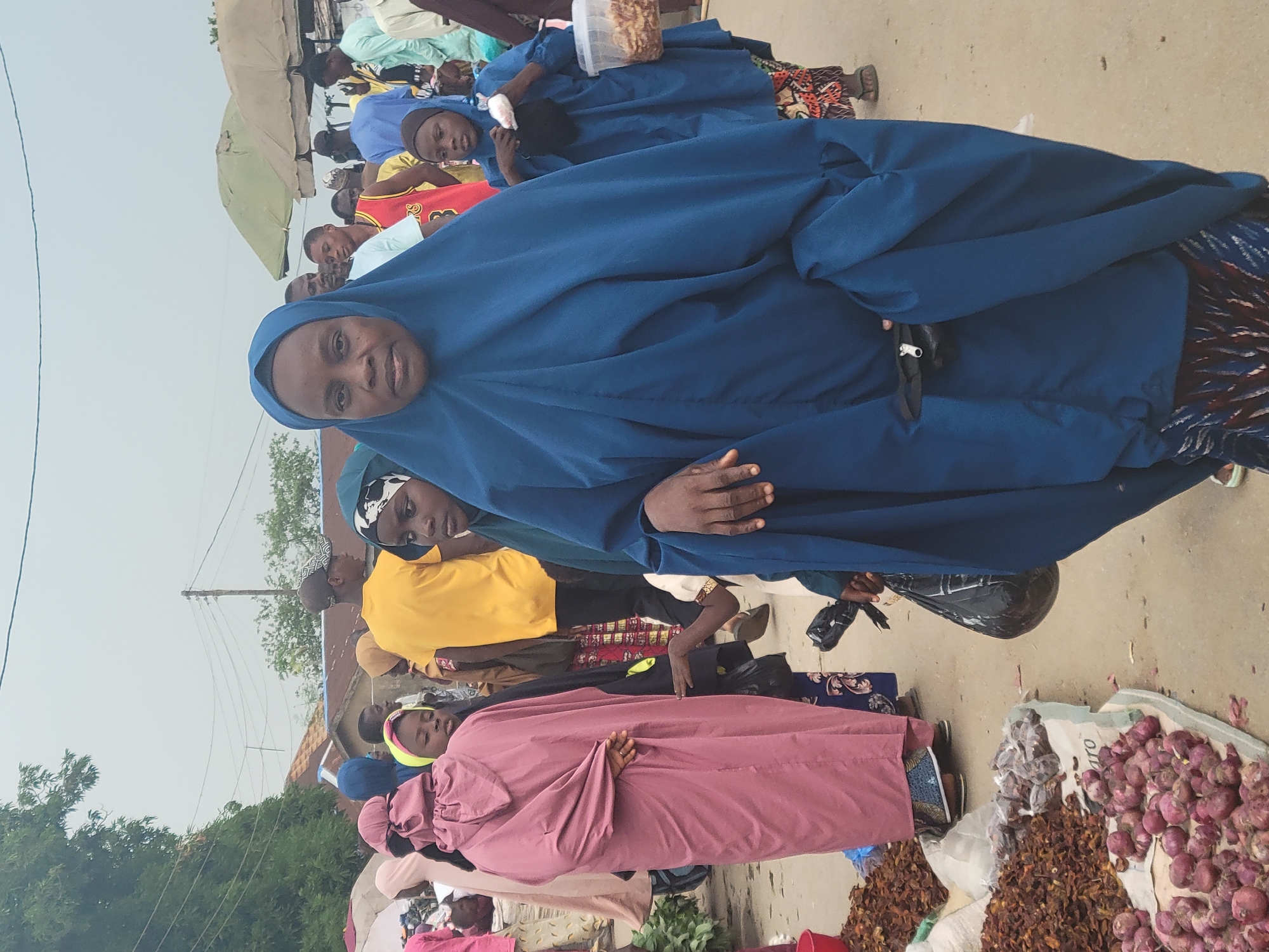 The image depicts a street scene in Madalla, Nigeria. Several people, primarily women and children, are visible. The women are wearing hijabs and traditional clothing in various colors, including blue and pink. Some individuals appear to be shopping at a market. Piles of onions and other dry goods are displayed on the ground. Other people can be seen in the background. The setting appears to be a bustling outdoor marketplace in broad daylight.