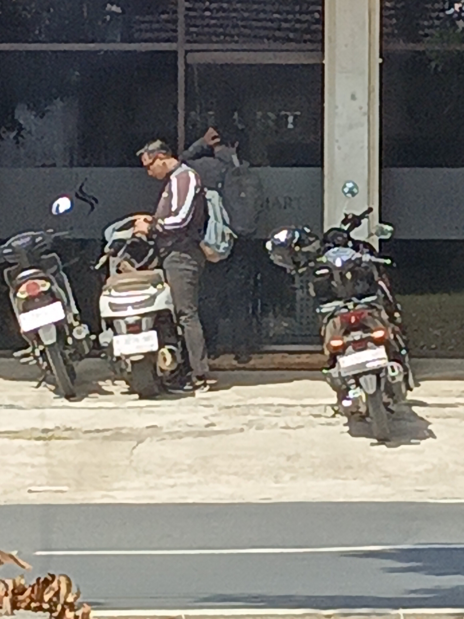 In Srono, Indonesia, a man is seen standing between two parked motorcycles. He is wearing a dark jacket with white stripes and dark pants. He appears to be adjusting something on the motorcycle in the center of the frame, which is white and has a light-colored seat. A blue and green backpack is slung over his shoulder. To his left, another motorcycle, predominantly black, is parked. To his right, a third motorcycle, also dark in color, is parked. The motorcycles are situated on a paved surface, likely a sidewalk or parking area, adjacent to a building with large glass windows. The lighting suggests it is daytime.