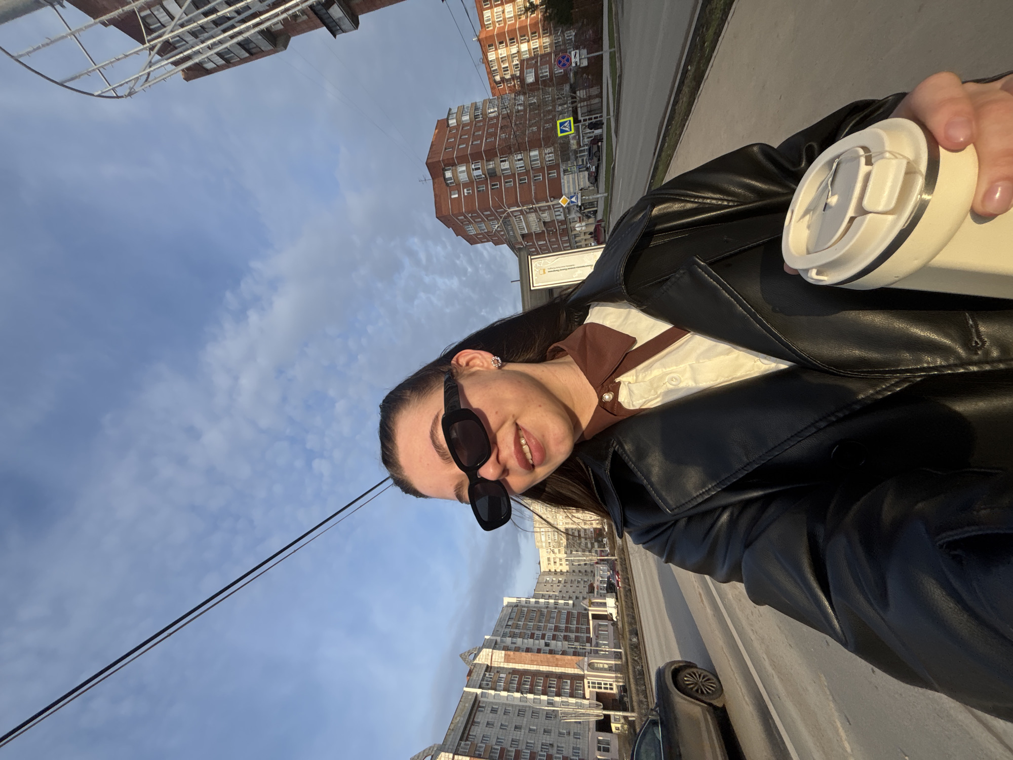 A woman stands in Perm, Russia, holding a white reusable coffee cup in her right hand. She is wearing a black leather jacket over a white collared shirt with a brown trim. Sunglasses frame her face, and she has dark hair pulled back. The background includes multistory brick buildings under a blue sky with scattered clouds. A street and parked cars are visible on her right. Power lines extend diagonally across the upper left of the frame. A pedestrian crossing sign is visible.