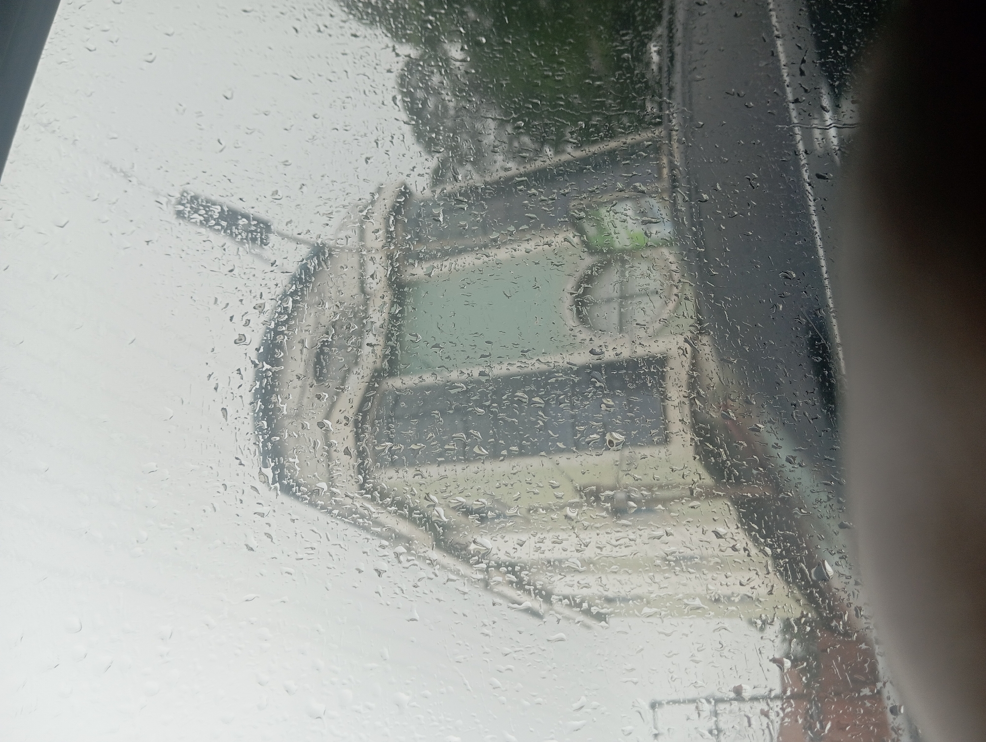 The image is taken through a rain-streaked car window, offering a blurred view of a building and its surroundings in Onitsha, Nigeria. The building appears to be light-colored with some architectural details, possibly a public institution or a commercial establishment, as suggested by the green signage visible through the rain. The sky is overcast and the weather is clearly inclement, as indicated by the numerous water droplets distorting the view. In the foreground, a portion of the car's dashboard or interior is visible, suggesting the photograph was taken from inside a vehicle. The scene is dominated by the muted colors and diffused light typical of a rainy day.