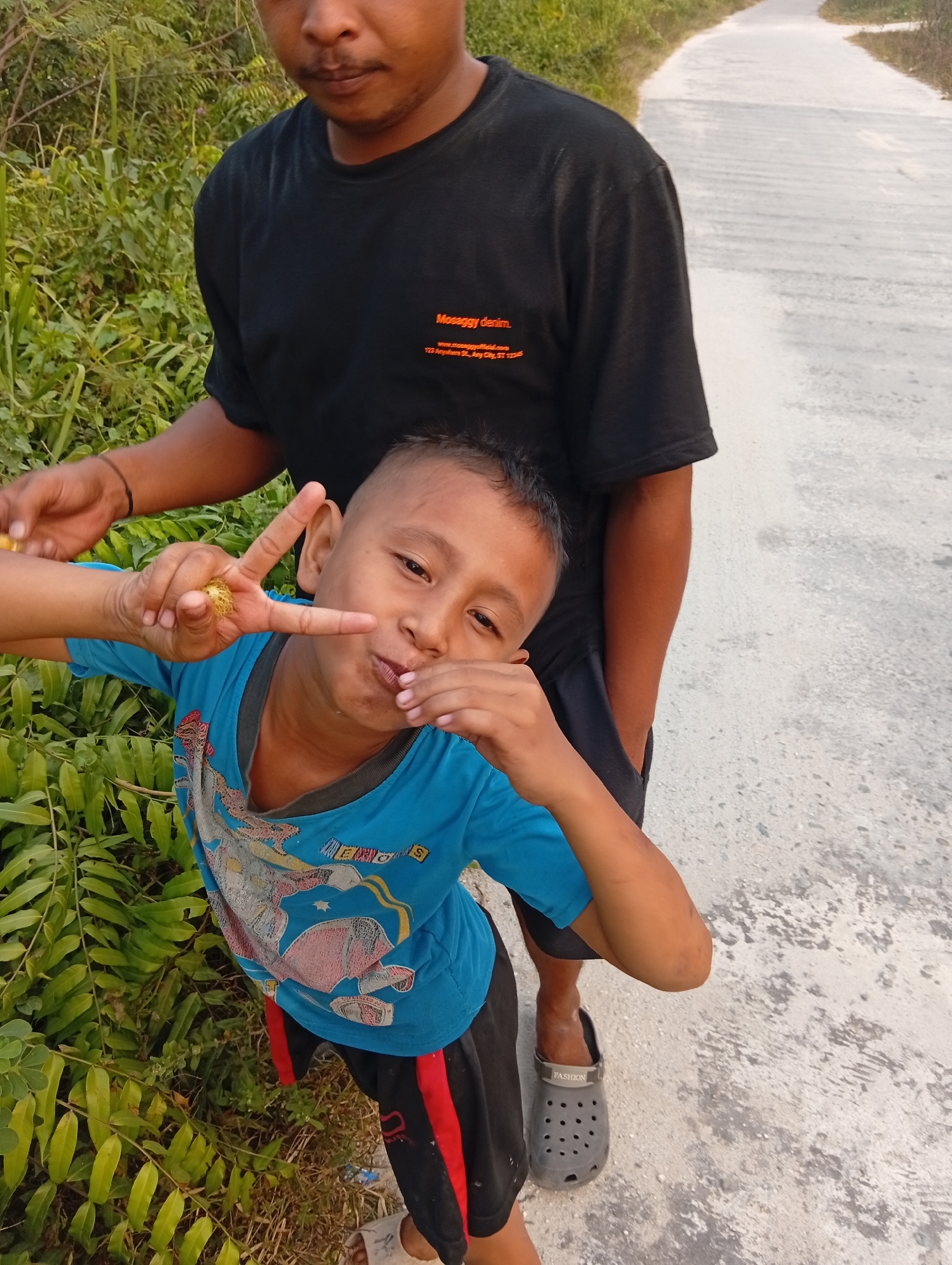 The image depicts two individuals, an adult and a child, positioned outdoors on a concrete path that appears to be a road in a rural or semi-rural setting. The adult, partially visible from the chest down, wears a black t-shirt with an orange text graphic that reads "Mosaggy Denim" along with a website and address. The child, in the foreground and facing the camera, is wearing a blue t-shirt with a graphic print and black shorts with red stripes. The child is making a "peace" sign with their right hand, and appears to be playfully putting a small, spiky object in their mouth with their left hand. The background consists of dense green vegetation on the left and the concrete path stretching into the distance on the right. The lighting suggests it is daytime, likely late afternoon given the warm light. The location is specified as Kualu, Indonesia.