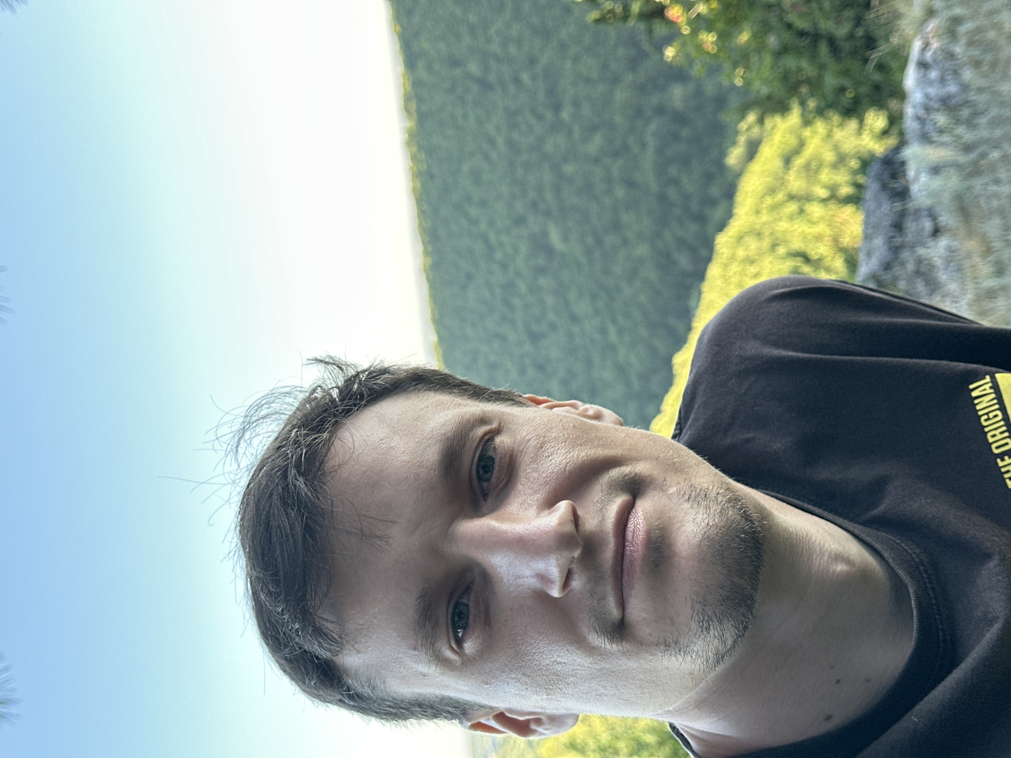 A man with short, dark hair and stubble is looking at the camera. He is wearing a dark t-shirt. He is outdoors, with a blurred background of green trees and a bright sky. The image appears to be a selfie, taken from a low angle. The man's face is in sharp focus, while the background is out of focus.