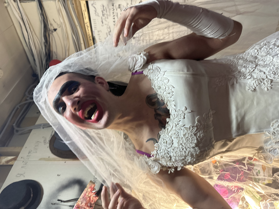 A person wearing heavy makeup, including exaggerated dark eyebrows and bright red lipstick, is dressed in a white wedding gown and veil. They have tattoos on their chest and arm, and wear white gloves. The individual is posed in a dramatic, expressive manner, with their mouth open. The background appears to be an interior space, possibly a storage or dressing room.