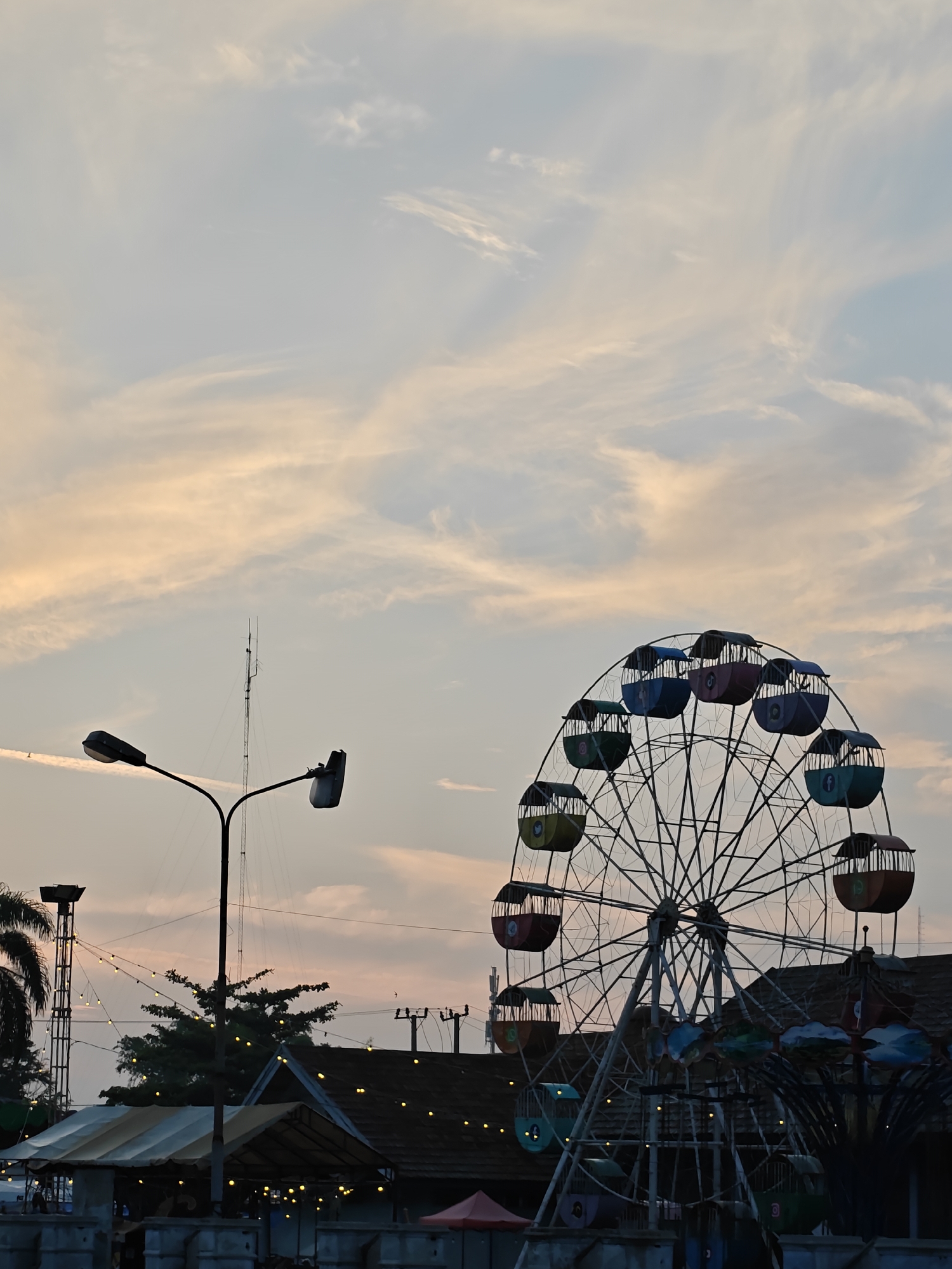 The image depicts an amusement park scene at dusk in Lhokseumawe, Indonesia. The sky is filled with soft, wispy clouds painted with hues of orange and pale blue, indicative of a sunset. Dominating the right side of the frame is a Ferris wheel with colorful gondolas, some displaying logos of social media platforms. To the left, a street lamp with two heads stands tall against the sky, with a communication tower visible in the background. Below the Ferris wheel and street lamp, a low-rise building with a tiled roof and a covered structure with string lights are visible, suggesting an area for visitors. The overall atmosphere is calm and recreational, characteristic of a public park or fairground in the city.