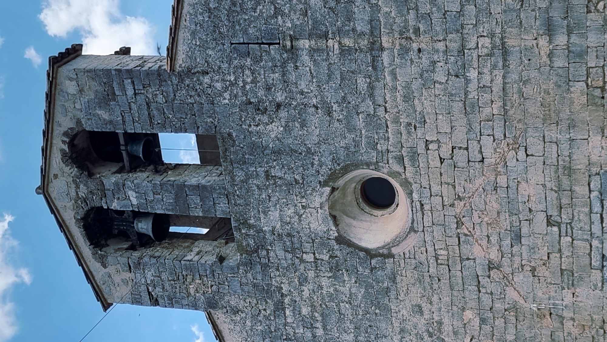 The image shows the facade of a stone building with a bell tower. The bell tower has two arched openings, each containing a bell. Below the bell tower is a circular window with a dark interior. The stone blocks of the building are weathered and uneven. The sky above is blue with some scattered clouds. The building is located in Montagut i Oix, Spain.