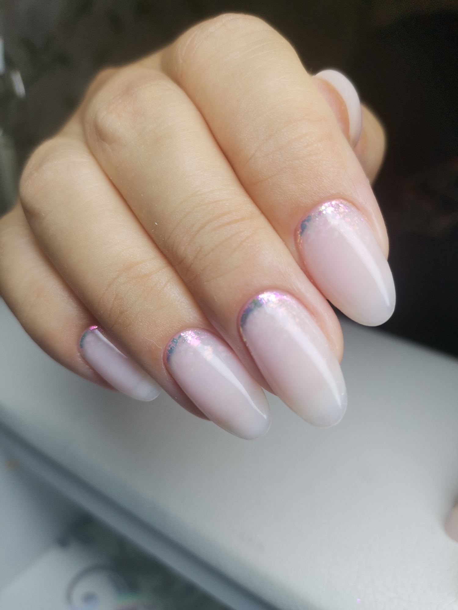 A close-up, eye-level shot focuses on a person's hand with manicured fingernails. The nails are almond-shaped and painted a translucent pale pink with a white ombré effect at the tips. Each nail features a subtle iridescent glitter gradient along the cuticle line, creating a delicate French manicure style. The skin of the hand appears smooth and fair, with visible finger creases. The background is softly blurred, showing hints of dark and light tones. There is no discernible information about the location in Kizel, Russia.