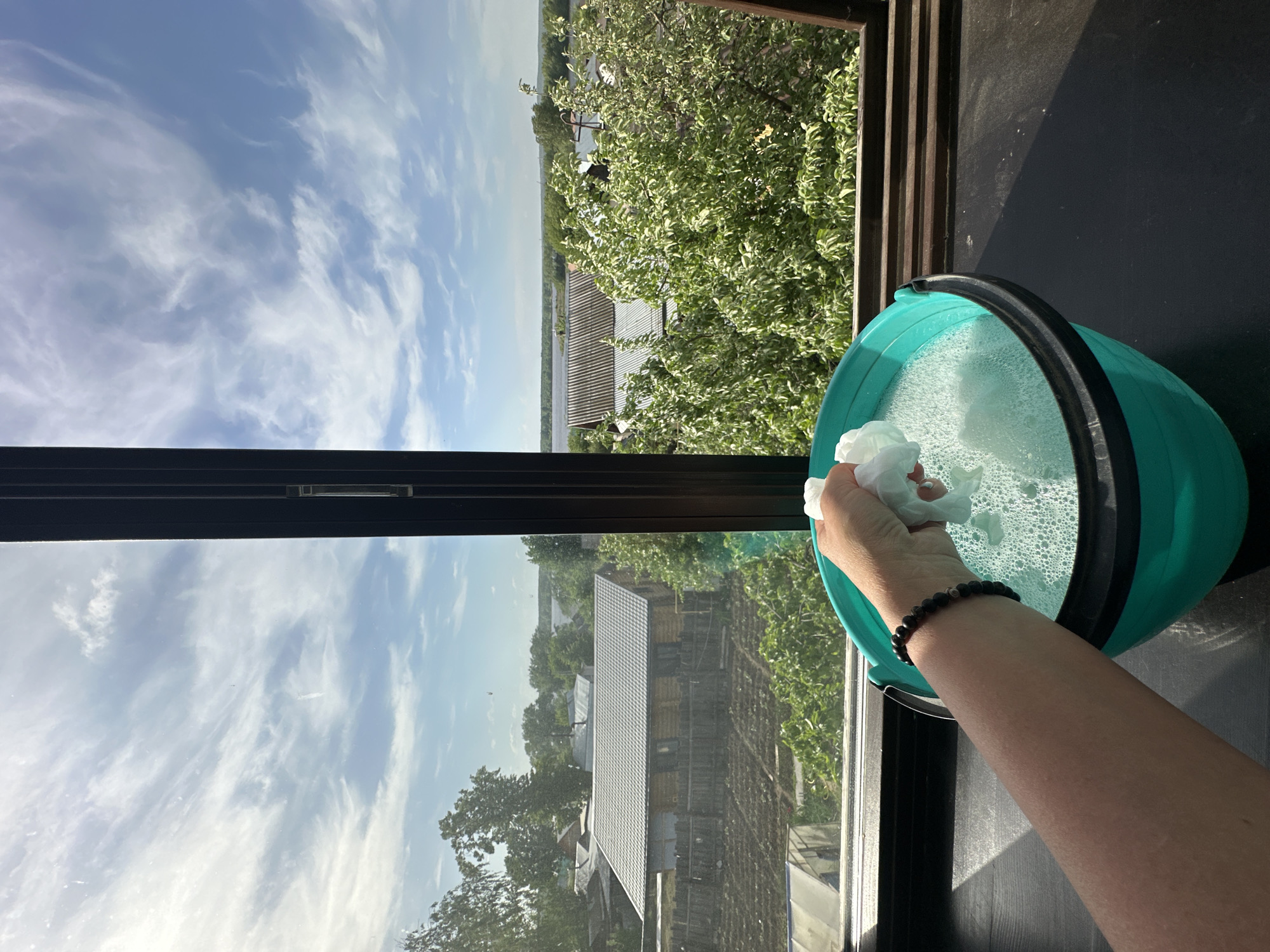 A person cleans a window in Chaikovsky, Russia. The camera is positioned indoors, looking out through a window with a dark frame. Visible outside is a blue sky with wispy white clouds, along with green trees and rooftops of buildings, suggesting a residential area. The person is holding a white cloth or sponge over a turquoise bucket filled with soapy water. A dark bracelet is visible on the person's wrist. The area immediately surrounding the bucket appears to be a dark, possibly wooden, surface.