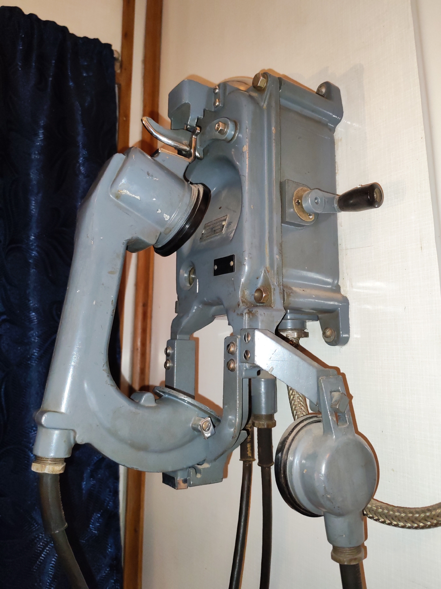 An old, grey, metal wall-mounted telephone is the primary subject of the image. It is mounted on a white wall, and a dark blue curtain hangs to the left of the phone. The phone has a hand crank on its right side and a receiver hanging on the left. The image does not depict any details regarding the location or its relation to the city of Bol'shie Mkhi, Russia, people, subjects, actions, or interactions.