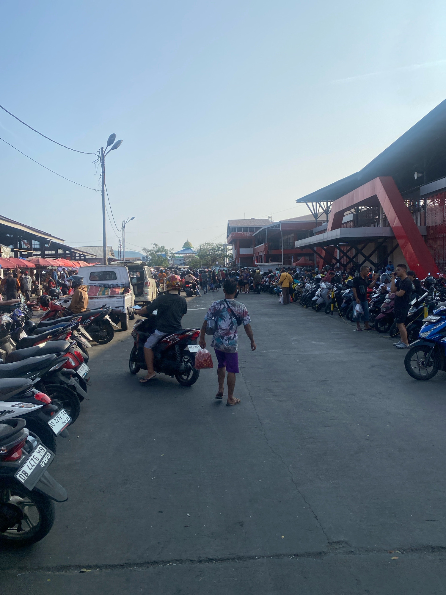 The image depicts a street scene in Manado, Indonesia, likely a market area. The foreground is dominated by a paved road with several parked motorcycles. Two individuals are in the process of riding motorcycles, one carrying a bag of produce. In the background, a bustling crowd of people is visible, along with market stalls and various buildings. The architecture suggests a modern commercial or public space with some red accents on the buildings. The sky is clear and bright, indicating daytime.