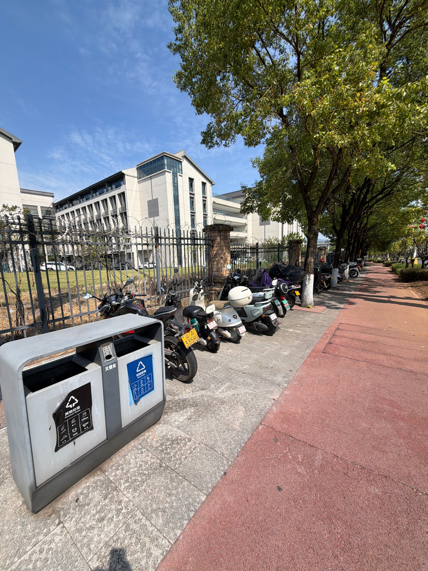 The image depicts a street scene in Jiangjin, China, on a clear day. In the foreground, a grey, dual-compartment trash receptacle is positioned on a paved sidewalk. One compartment is labeled with Chinese characters and a recycling symbol, indicating it is for recyclables. The other compartment has a black sticker with Chinese characters and pictograms depicting items like a banana peel and a crumpled piece of paper, labeled as "Other Waste." Beyond the trash can, a row of parked motorcycles and scooters lines the sidewalk. A black metal fence with barbed wire runs along the edge of the property, separating the sidewalk from a grassy area and a modern, multi-story building in the background. The building has a light-colored facade with numerous windows and balconies. Trees with green foliage are planted along the sidewalk, casting shadows on the ground. The sidewalk transitions to a reddish-colored path to the right of the frame, suggesting a pedestrian walkway. The sky is bright blue with scattered white clouds. No people are visible in the scene.