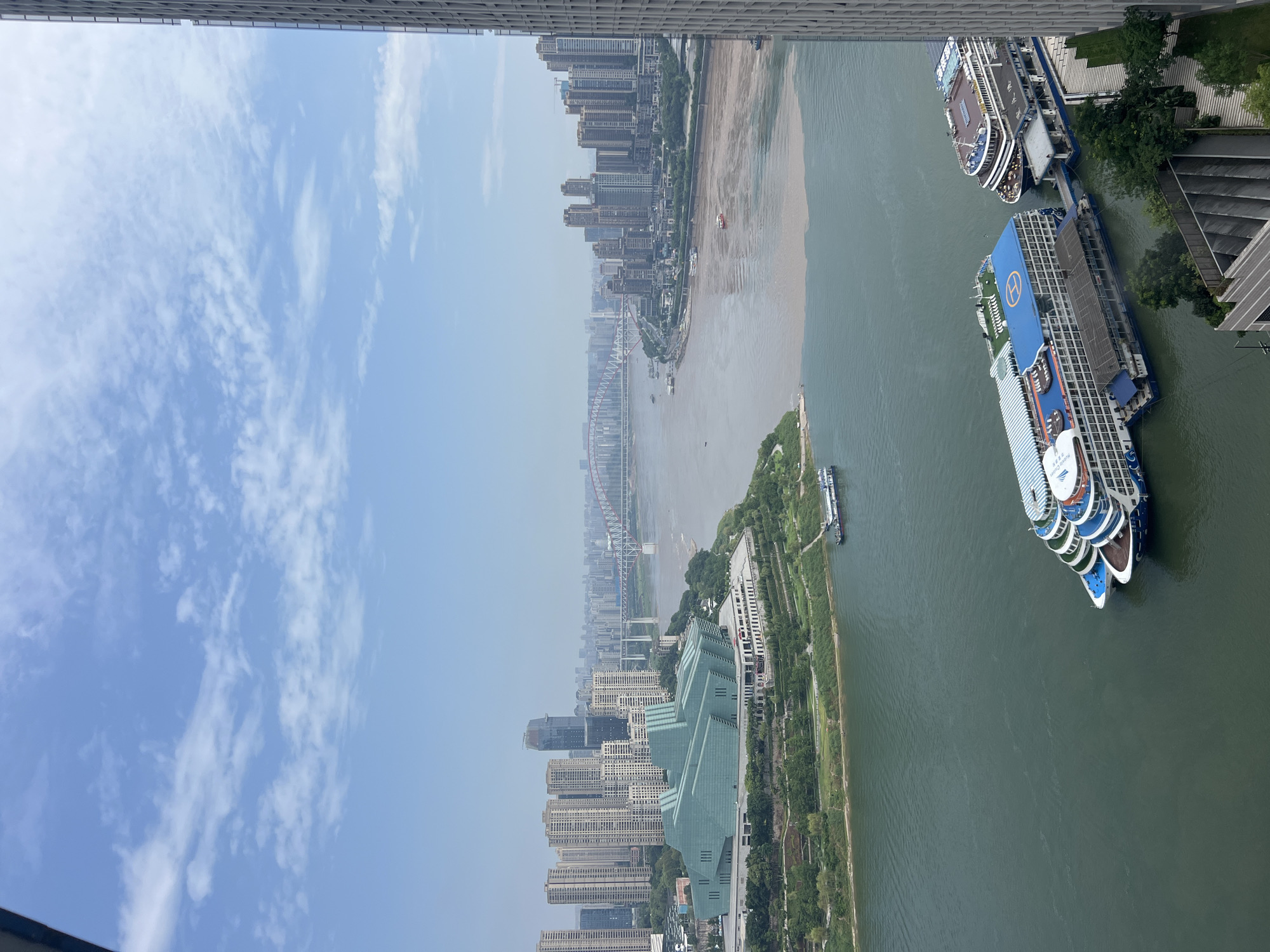 The image captures a panoramic view of the Yuzhong District in Chongqing, China, focusing on the confluence of the Yangtze and Jialing Rivers under a hazy sky with scattered clouds. In the foreground, three large, white cruise ships with blue accents are docked. The largest ship features a prominent blue deck with a helipad marked with a blue "H". Adjacent to it, another ship of similar design is connected, and further to the right, a larger, darker ship with multiple decks is docked. These vessels are positioned on the calmer, greener waters of one of the rivers.
The middle ground is dominated by the meeting of two rivers, one appearing a murky brown and the other a clearer green, creating a distinct line where they converge. Several smaller boats navigate the waterways. On the left bank, a distinctive, angular green building with a faceted facade stands prominently amidst lush green terraced landscaping and trees. Behind this building, numerous tall residential skyscrapers rise against the skyline.
Further in the distance, the cityscape unfolds with a dense cluster of modern buildings and skyscrapers. A large, red suspension bridge spans the wider river, connecting different parts of the city. The horizon is dotted with more buildings and a hazy atmosphere characteristic of urban environments. The right edge of the image is framed by the side of a tall, modern building with a grid-like pattern of windows. The overall scene depicts a bustling urban landscape at the confluence of major rivers, highlighting modern architecture and riverine transportation.