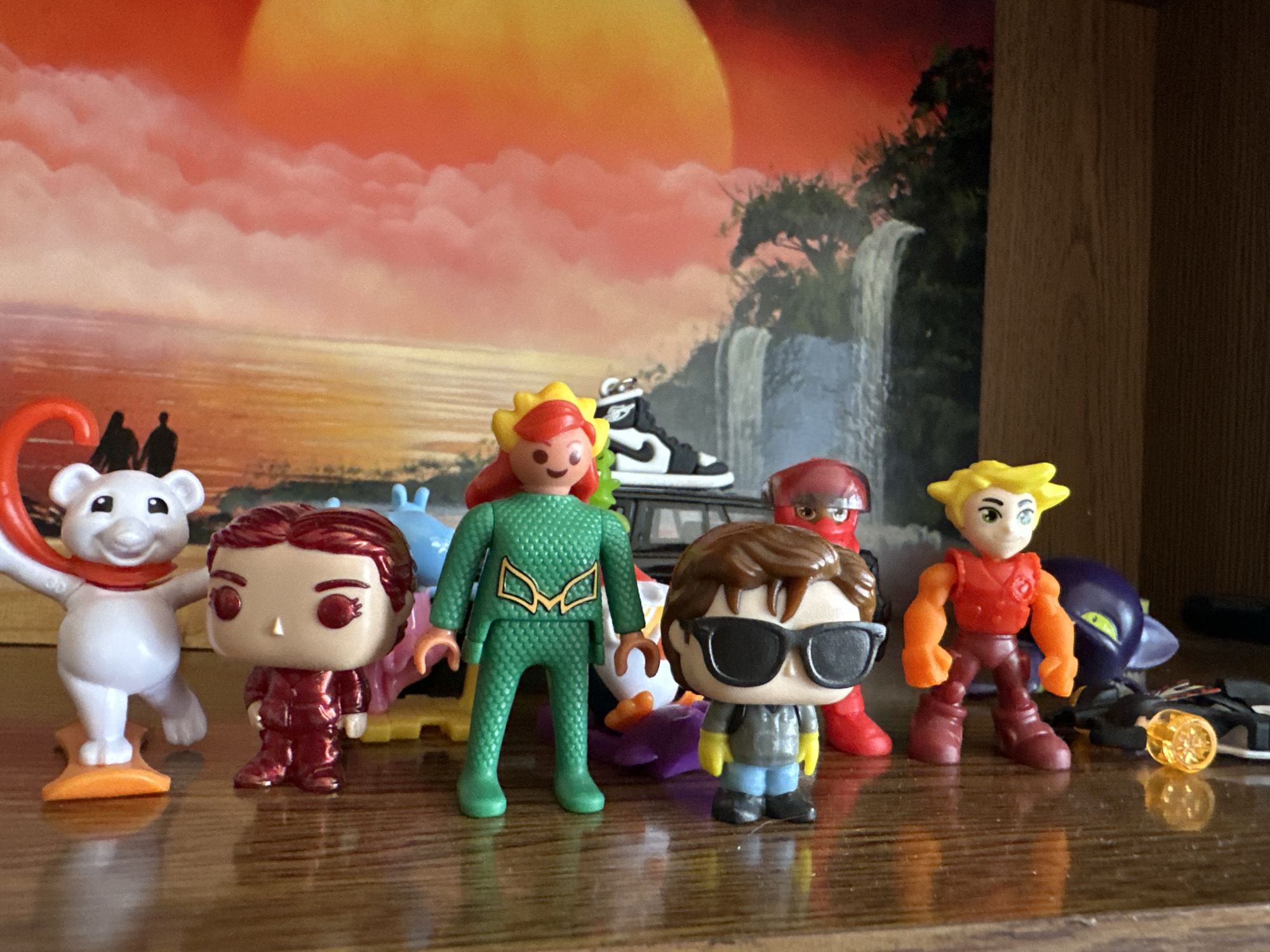In an indoor setting, likely a shelf, a collection of action figures are displayed. The figures include a white polar bear with a red scarf, a metallic red Funko Pop figure, a green-clad figure with yellow hair, a figure wearing sunglasses and a grey jacket, a red figure with yellow hair and orange arms, and a purple dragon-like figure. In the background, a poster or artwork depicts a sunset over water with silhouettes of two people and a waterfall scene. The figures are arranged on a wooden surface.