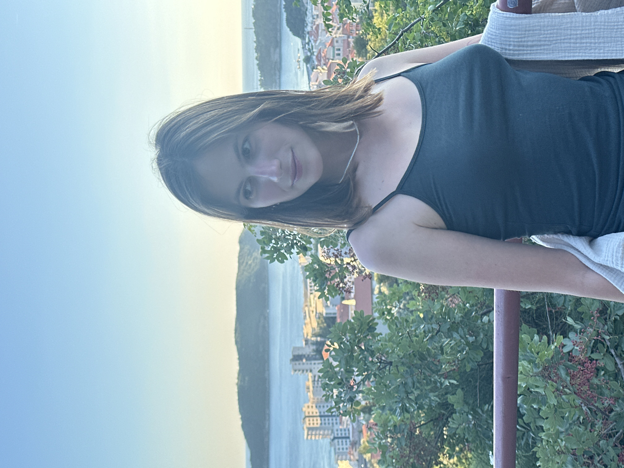 A young woman stands on a balcony overlooking the city of Čučuci, Montenegro. She has shoulder-length brown hair and is wearing a dark green camisole and a thin necklace. She is holding a light-colored blanket over her arm. The background shows a coastal city with buildings and a body of water, with hills in the distance under a clear blue sky. Lush green foliage is visible in the foreground and around the balcony. The lighting suggests it is late afternoon or early evening.