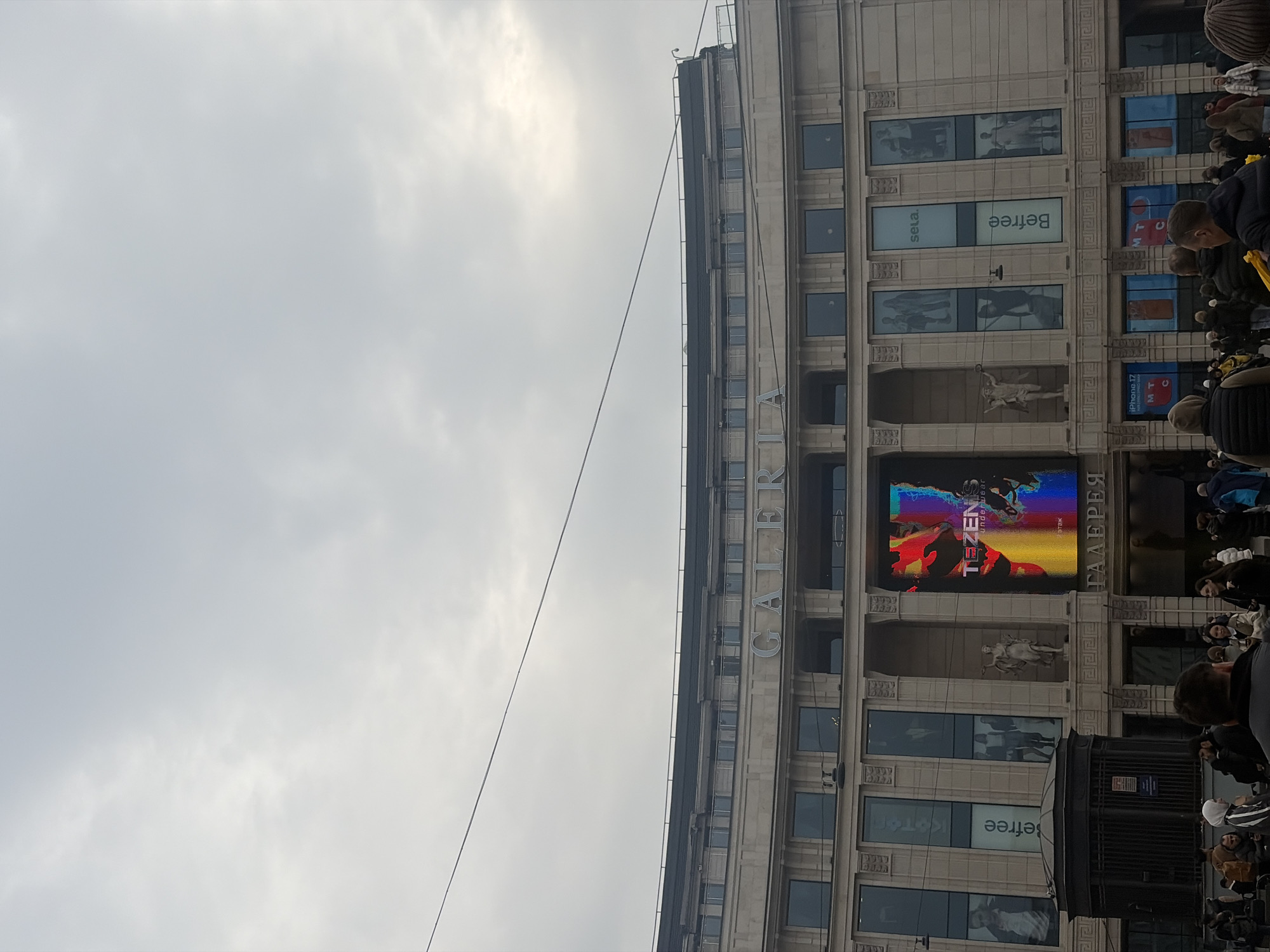 This image depicts the exterior of the Galeria Shopping Mall, located at Ligovsky Prospekt 30A in Saint Petersburg, Russia. The scene is observed under an overcast, light gray sky.
The prominent central subject is the multi-story, light-colored stone facade of the mall, featuring classical architectural elements. The word "GALERIA" is displayed in large letters on the upper section of the building. A large vertical LED screen is centrally located on the facade, displaying an abstract, multi-colored graphic and the text "TEZENIS UNDERWEAR." Below this screen, the Russian word "ГАЛЕРЕЯ" is also visible. Flanking the LED screen are vertical window displays featuring advertisements for "Befree" and "sela." Two classical statues are situated in niches on either side of the main LED display. Lower-level windows display imagery related to "iPhone 17" and the "MTS" (МТС) telecommunications company.
In the foreground, a substantial crowd of pedestrians, dressed in various cold-weather garments such as jackets and hats, occupies the street level. Most individuals are walking or standing, with their backs or sides to the camera. To the left, a dark, octagonal kiosk-like structure with vertical railings is visible, suggesting an entrance to an underground passage or a metro station. A single, thin overhead utility wire traverses the upper-mid section of the frame horizontally. No specific interactions or detailed actions are discernible within the crowd.