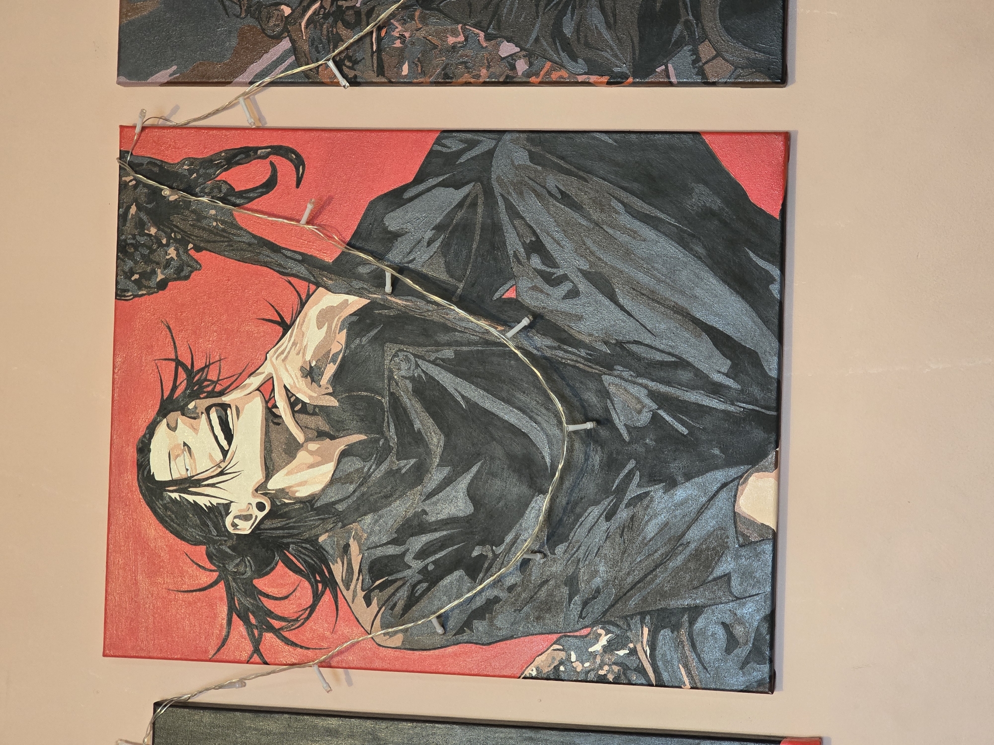 The image shows a framed painting of a man with long black hair tied in a bun. He has his eyes closed and a wide smile, with his right index finger touching his chin. He is wearing a dark outfit with intricate folds. A dark, abstract object with claw-like protrusions is visible to his right. The painting has a red background and is adorned with a string of small, white LED lights. The painting is displayed on a light pink wall, and portions of two other similar paintings are visible on either side.