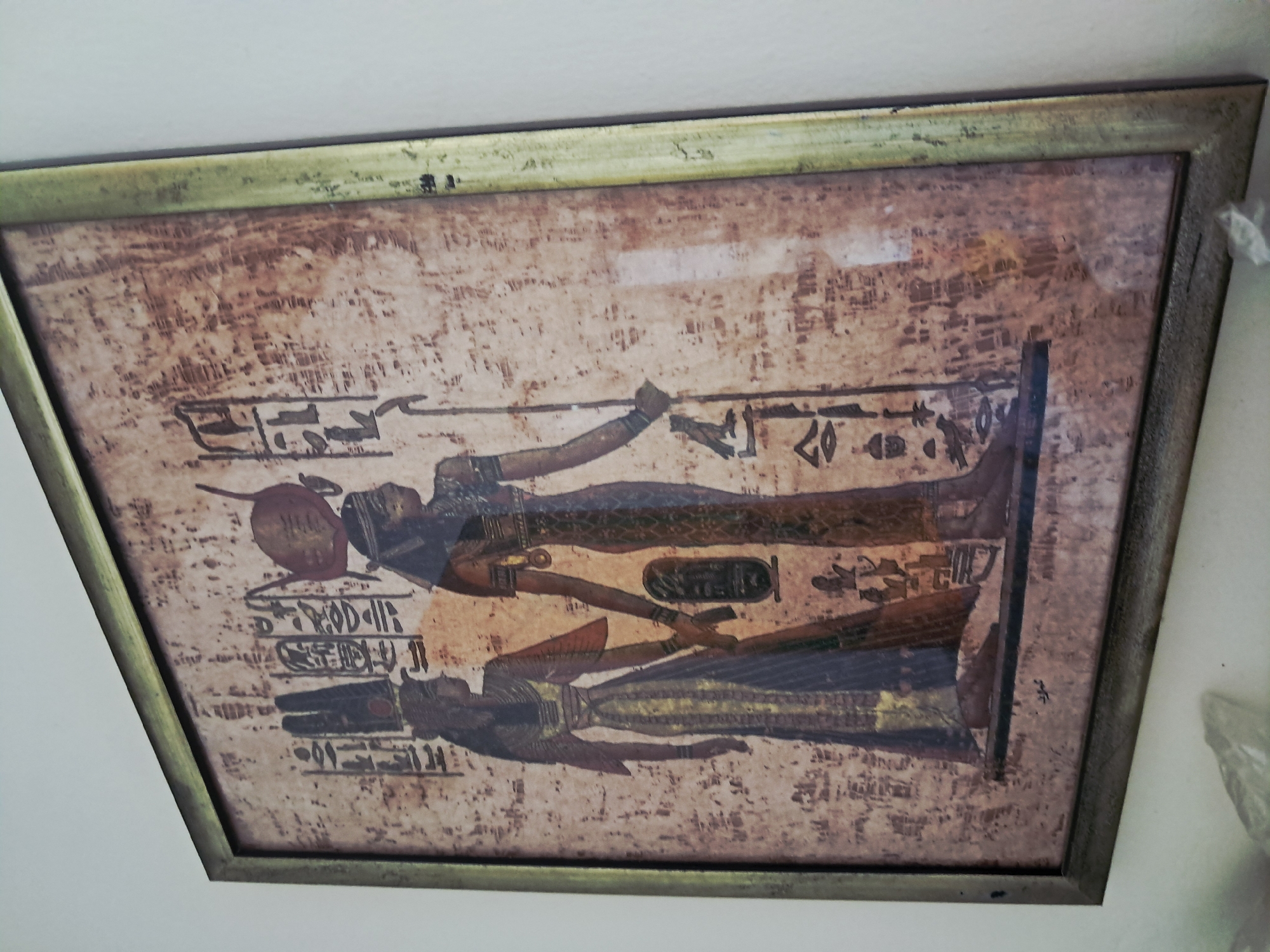 A framed artwork depicting an Egyptian-style scene is mounted on a white wall. The artwork features two figures in profile, adorned in what appears to be traditional Egyptian garb, surrounded by hieroglyphs on a textured, aged background resembling papyrus. The frame is metallic with a tarnished gold or bronze finish. The picture is hung slightly skewed.
There is no information related to Juan Griego, Venezuela in the image or the surrounding context.