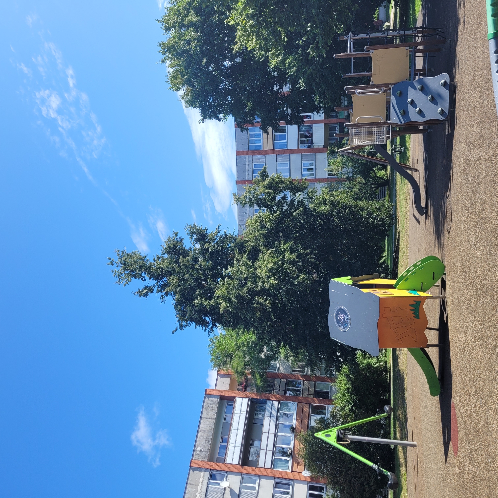 This image shows a playground with children's play equipment in the city of Jūrmala, Latvia. The playground is located in front of several multi-story apartment buildings. In the foreground, there is a bright orange and green playhouse with a slide, and a green swing set with black seats. Further back, there is a larger play structure with slides, climbing walls, and nets. Tall, leafy trees are scattered throughout the scene, and the sky is a clear blue with a few wispy clouds. The ground of the playground is a soft, brown, rubberized surface. The buildings in the background appear to be of Soviet-era architecture, with balconies and numerous windows.