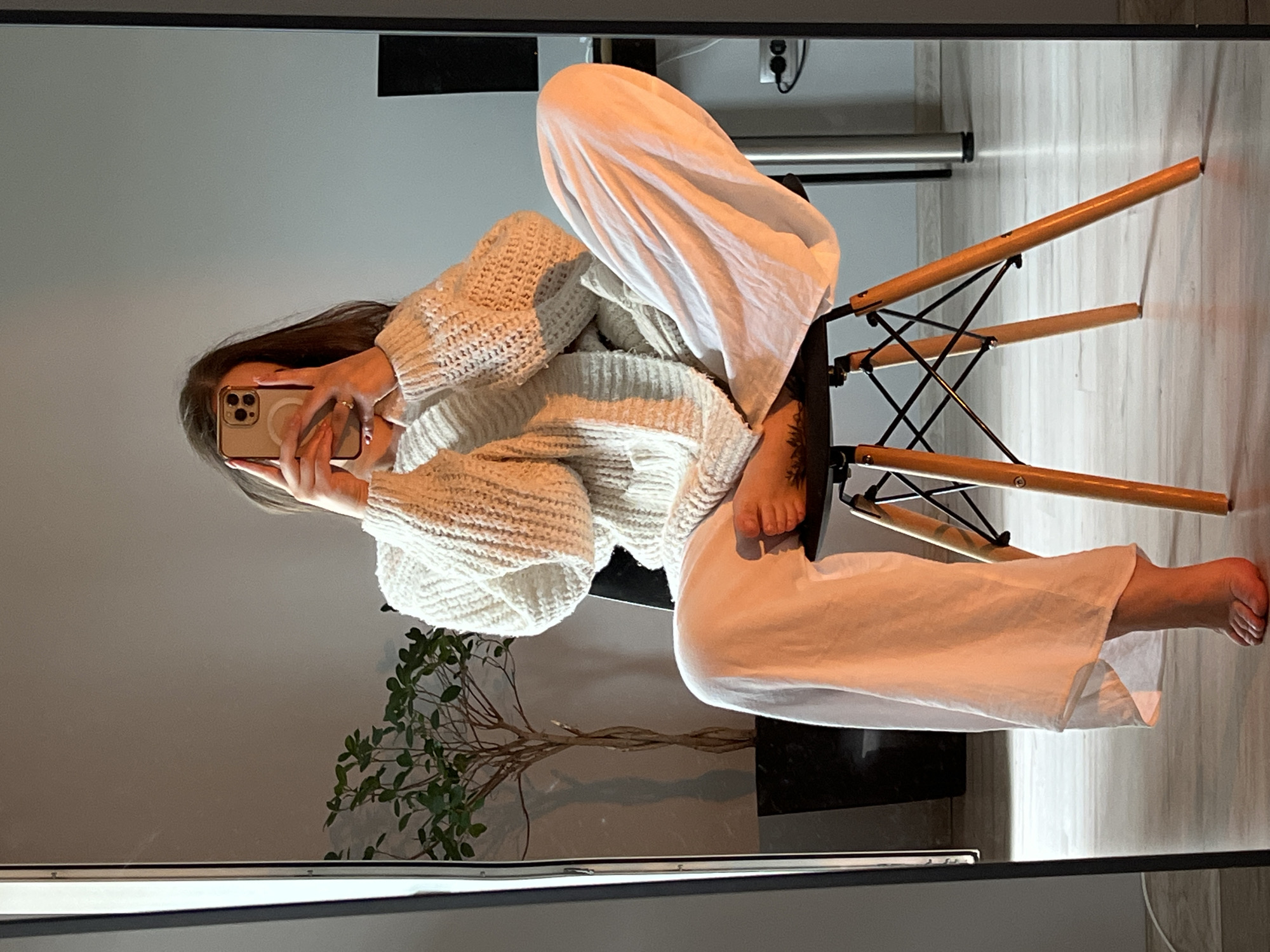 This image is a mirror selfie of a young woman captured indoors, presented with a 90-degree clockwise rotation. The woman, with long, dark brown hair, is seated in a relaxed, almost curled-up posture on a modern chair. She is wearing a cozy, light-colored (likely white or off-white) chunky knit sweater with voluminous sleeves and matching wide-leg pants. Her face is largely obscured by the light-colored smartphone she holds with both hands to take the picture; the phone displays multiple camera lenses and a circular attachment on its back. Her bare feet are visible, and a small tattoo can be seen on her left ankle/foot.
The chair she sits on features a black metal frame and wooden components, indicative of a contemporary design. The background reflected in the mirror reveals simple, light-colored walls, what appears to be a light wooden floor, and some electrical outlets. To her left, a small, leafy green plant with distinctive, dried-looking stems rests on a dark, reflective surface. The overall setting is a modern and uncluttered indoor environment. The lighting is soft and ambient, suggesting either daylight or well-distributed artificial light. The scene conveys a casual and comfortable atmosphere. No text is visible in the image. The location context is Ternopil, Ukraine.