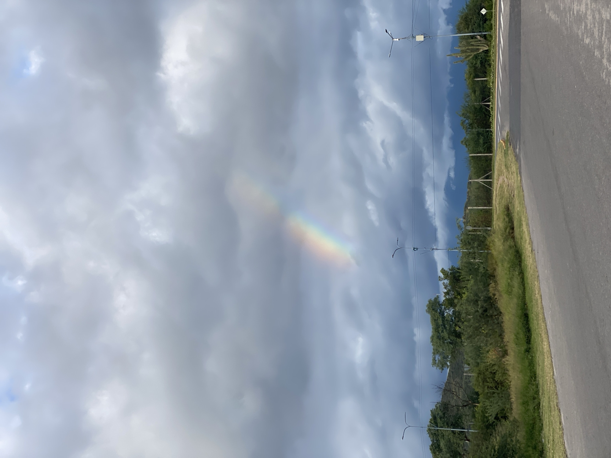 The image shows a landscape featuring a section of asphalt road on the right. Adjacent to the road is a grassy verge with trees and foliage. In the background, there are power lines and light poles extending into the distance. Above, the sky is filled with gray and white clouds, with a segment of a rainbow visible through the clouds.
While there is no precise location information from the image to find in San Fernando del Valle de Catamarca, Argentina, the scene depicts a road with green areas and utility poles near an area with likely open or natural spaces.