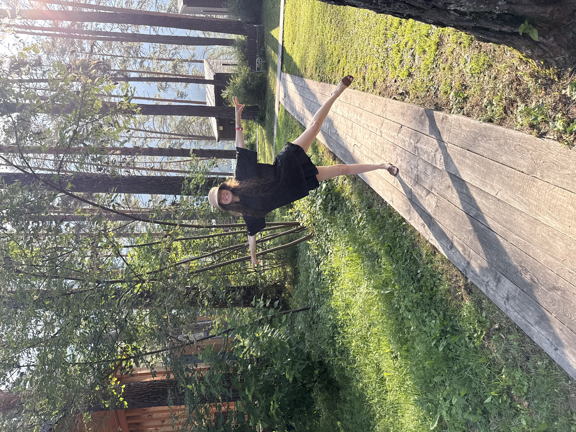In Barangol, Russia, a woman with long, wavy brown hair and a straw hat stands on a wooden boardwalk. She is wearing a black t-shirt dress and brown sandals. Her right leg is extended backward, and her arms are outstretched to the sides. She is smiling at the camera. The boardwalk is surrounded by lush green grass and tall trees, with wooden cabins visible in the background. The sun is shining, casting long shadows across the boardwalk.