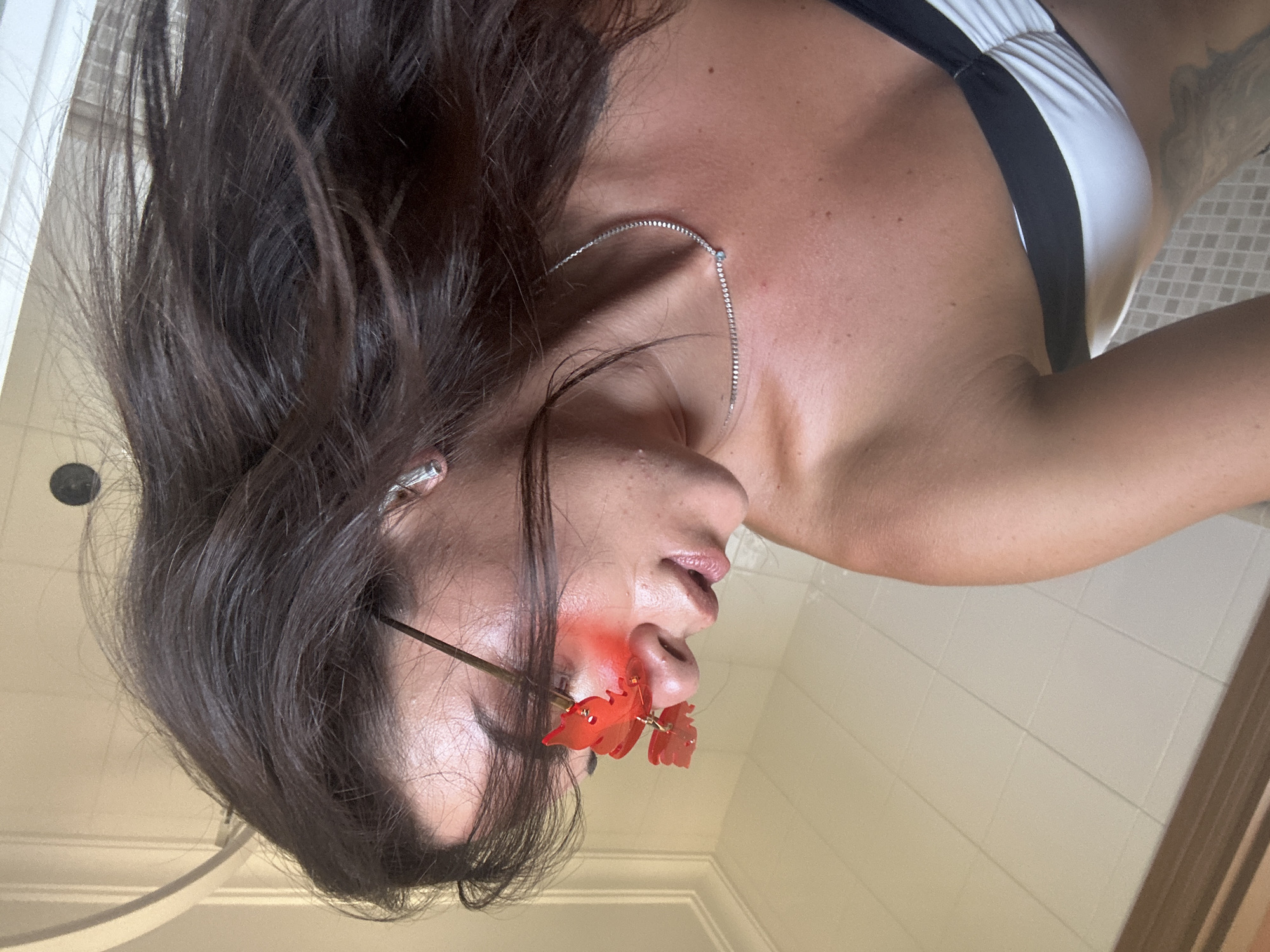 A person is captured in a close-up shot, angled upwards from the chest, in what appears to be a bathroom setting. The individual has dark brown hair that is slightly disheveled, framing their face. They are wearing distinctive red, butterfly-shaped sunglasses with a gradient lens effect that casts a reddish hue on their skin. A fine, silver necklace with a single light blue stone is visible around their neck. They are also wearing a bikini top that is predominantly white with black accents. The background shows light-colored tiled walls, and a white curved ceiling feature is visible at the top of the frame. A shower curtain or door is partially visible on the right side of the image. The overall lighting suggests an indoor environment. The location is identified as Capdepera, Spain, implying this bathroom is situated within the city.
