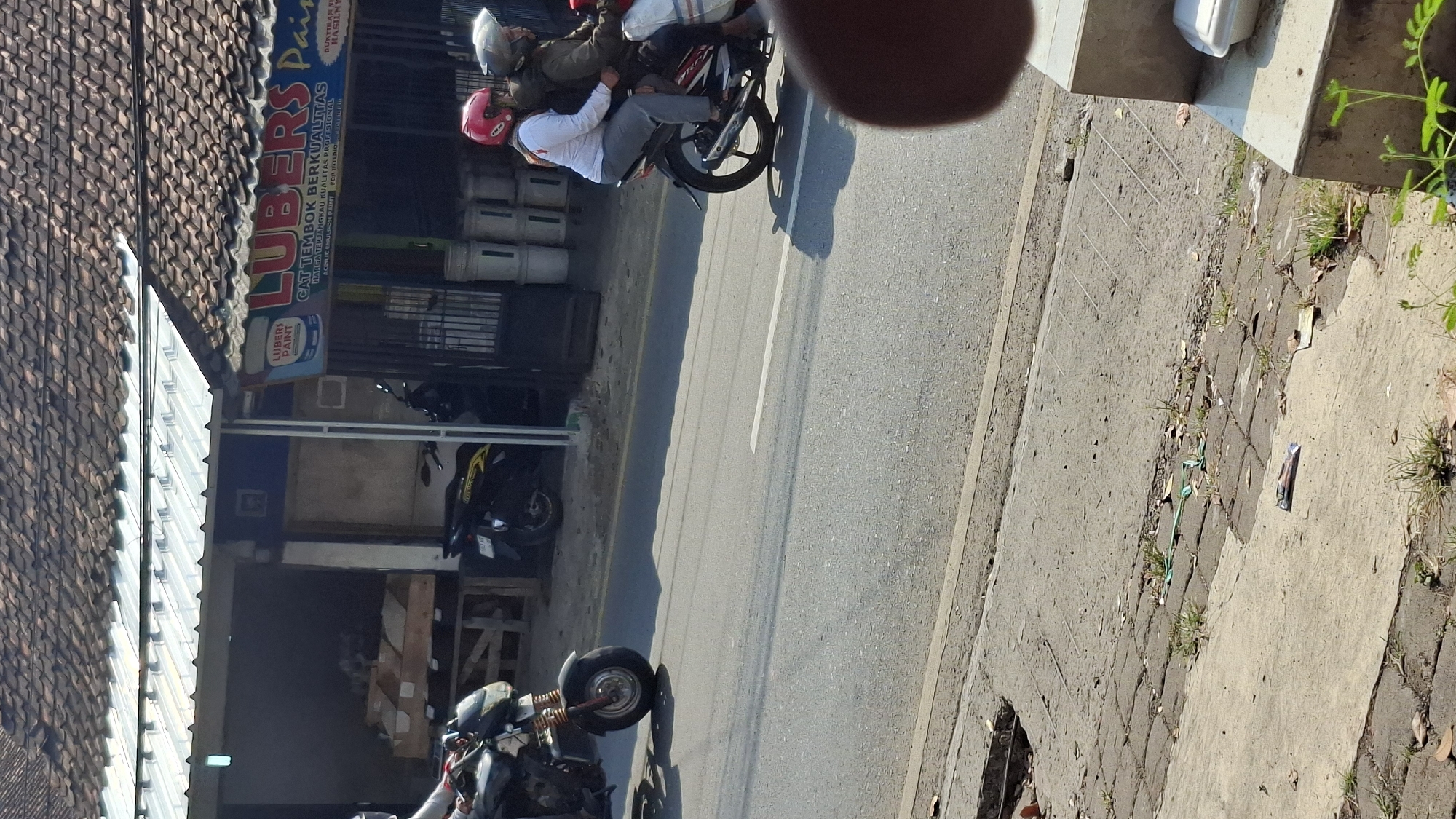 In Cilaku, Indonesia, on a sunlit day, two people ride a motorcycle down a paved road. The rider in front wears a green jacket and a gray helmet, while the passenger behind wears a pink helmet and a white long-sleeved shirt and gray pants. They are on a red and black motorcycle, moving away from the viewer. To their right, a row of shops lines the street. One shop prominently displays a large sign for "LUBERS PAINT" and advertises "CAT TEMBOK BERKUALITAS" (Quality Wall Paint) with smaller text mentioning "HARGA TERJANGKAU KUALITAS PROFESIONAL" (Affordable Price Professional Quality) and "ACRYLIC EMULSION PAINT FOR INTERIOR & EXTERIOR". Stacks of white paint buckets are visible behind the shop counter. Another motorcycle is parked in front of one of the shops. The road has white lane markings. In the foreground, a concrete and brick pathway runs alongside the road, with some scattered leaves and green plastic ties on the ground. A white foam food container sits on a concrete ledge in the lower right corner of the frame.