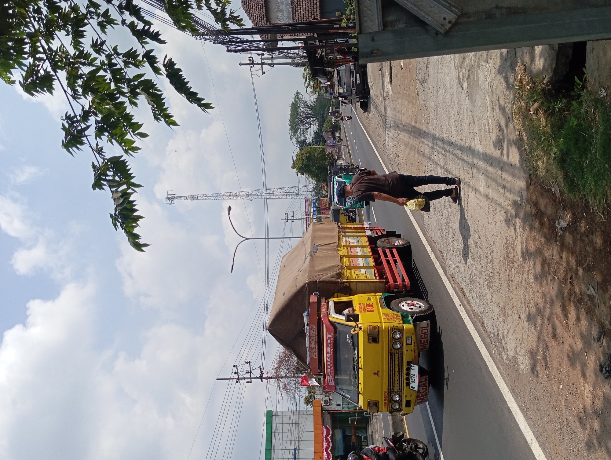 An eye-level, medium shot captures a sunny daytime scene on a paved road in Lemahbang, Indonesia. A bright yellow truck with a tan tarp covering its cargo is the primary subject, moving along the right lane of the road. The truck features red accents on its side panels and chassis. A person is walking away from the camera on the sidewalk to the right of the road, carrying a yellow plastic bag. The sidewalk is bordered by a grassy embankment and a concrete structure on the far right. Power lines and a tall communication tower are visible in the background, along with other vehicles and buildings suggesting a semi-urban or commercial area. The sky is partly cloudy, with lush green trees framing the top right portion of the image.