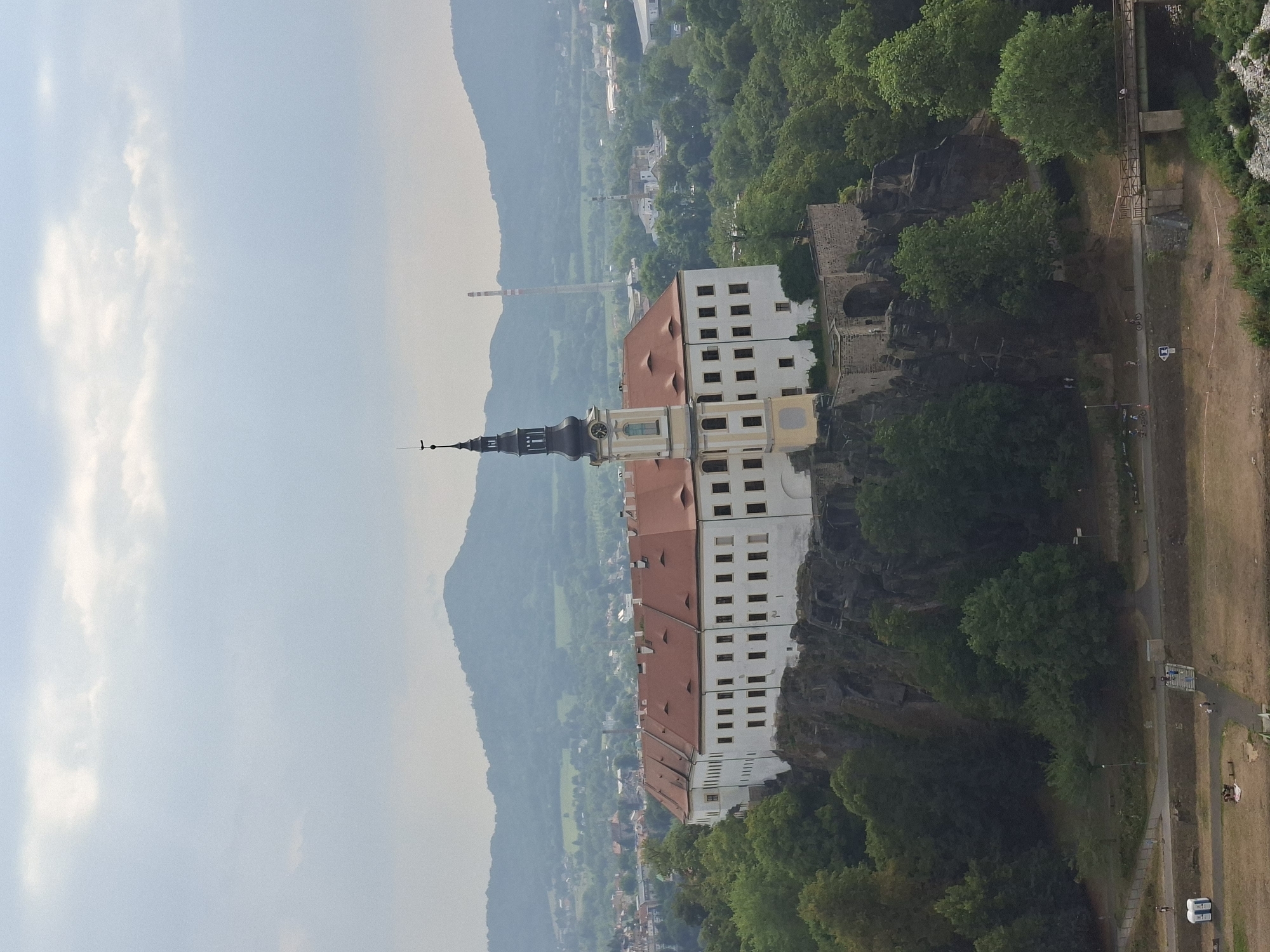 An aerial view of the Děčín Castle, located in Děčín, Czechia, shows the fortress perched on a rocky outcrop. The castle features a long white building with a red-tiled roof and a prominent black tower with a clock. Surrounding the castle is dense green foliage and a distant mountainous landscape under a cloudy sky. Below the castle, a pathway with a bridge crosses a body of water, and there are a few people visible walking or cycling. Two portable toilets are also present in the foreground. The scene captures the historical architecture of the castle amidst a natural and urban environment.